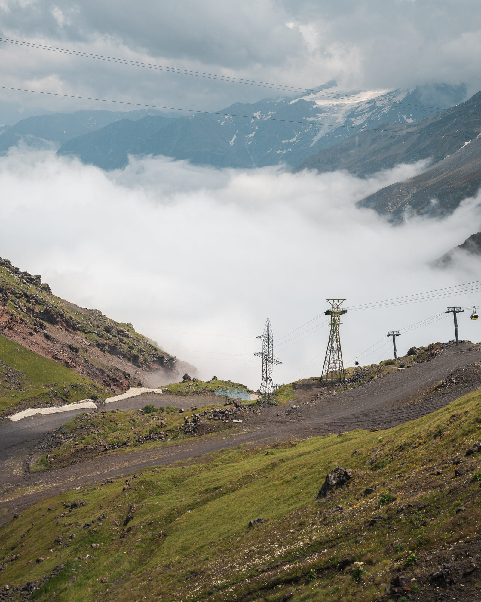 Cable car towers climbing into the clouds below Elbrus with road switchbacks on a green hillside