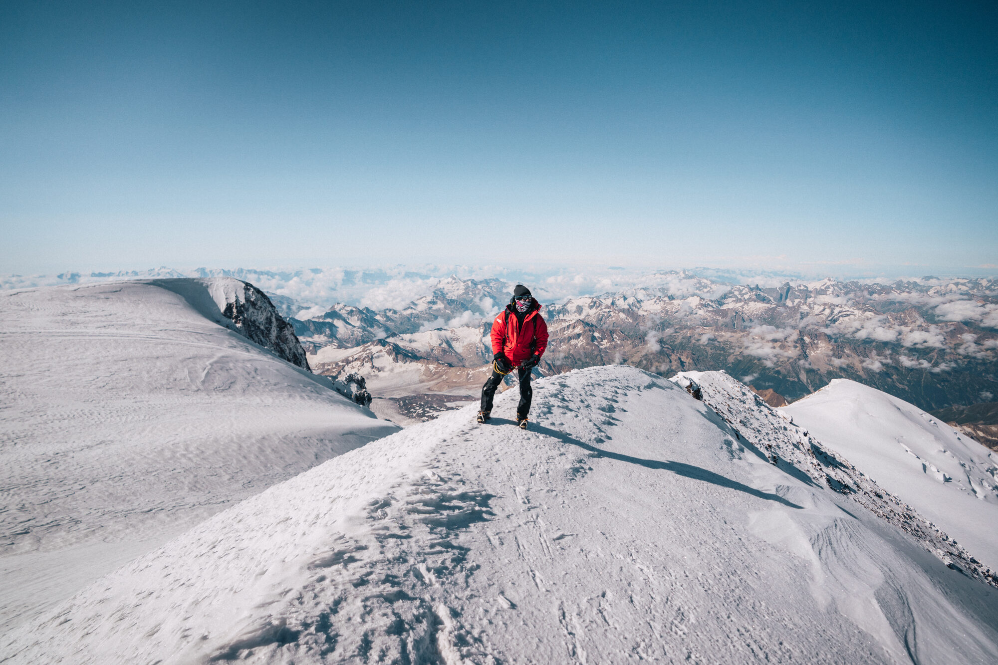 Climber in a red jacket walking along the narrow snowy summit ridge with the Caucasus behind