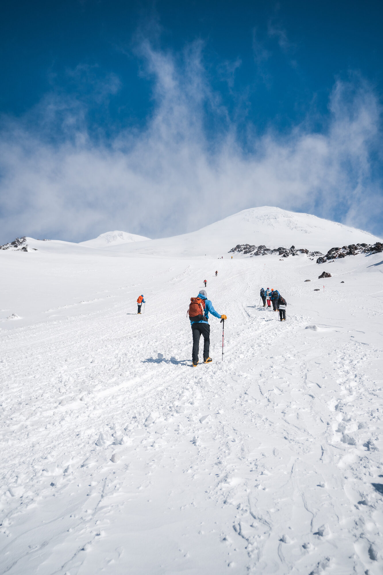 Climbers ascending a wide snow slope toward the twin summit cone of Elbrus under blue sky