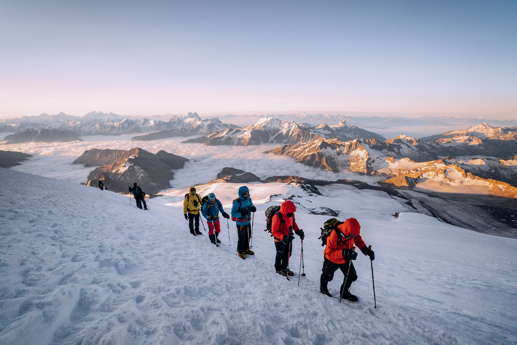 Six climbers in single file traversing the upper snow ridge of Elbrus at sunrise with the Caucasus lit gold below