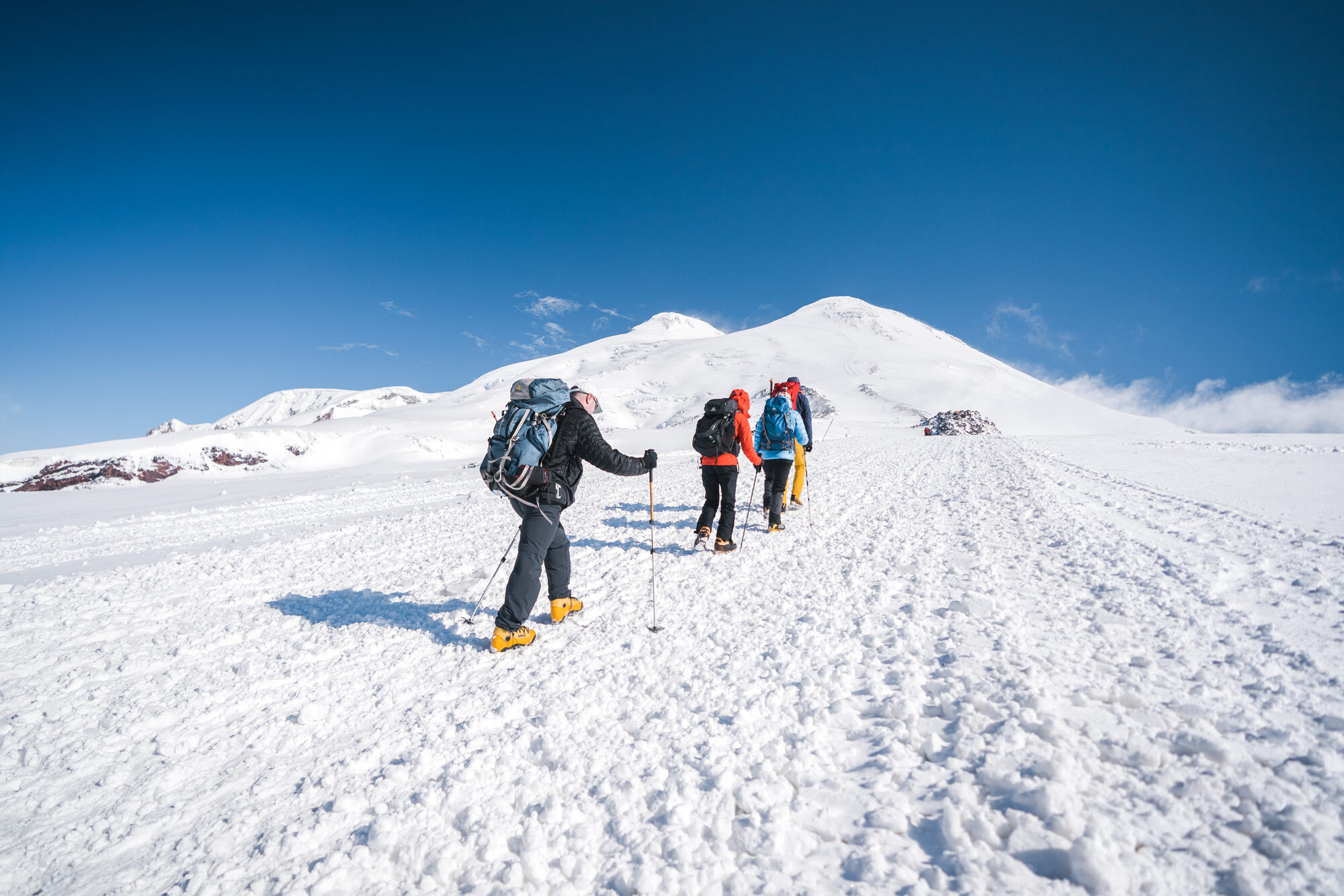 Four climbers ascending the snow field toward the twin summits of Elbrus under blue sky