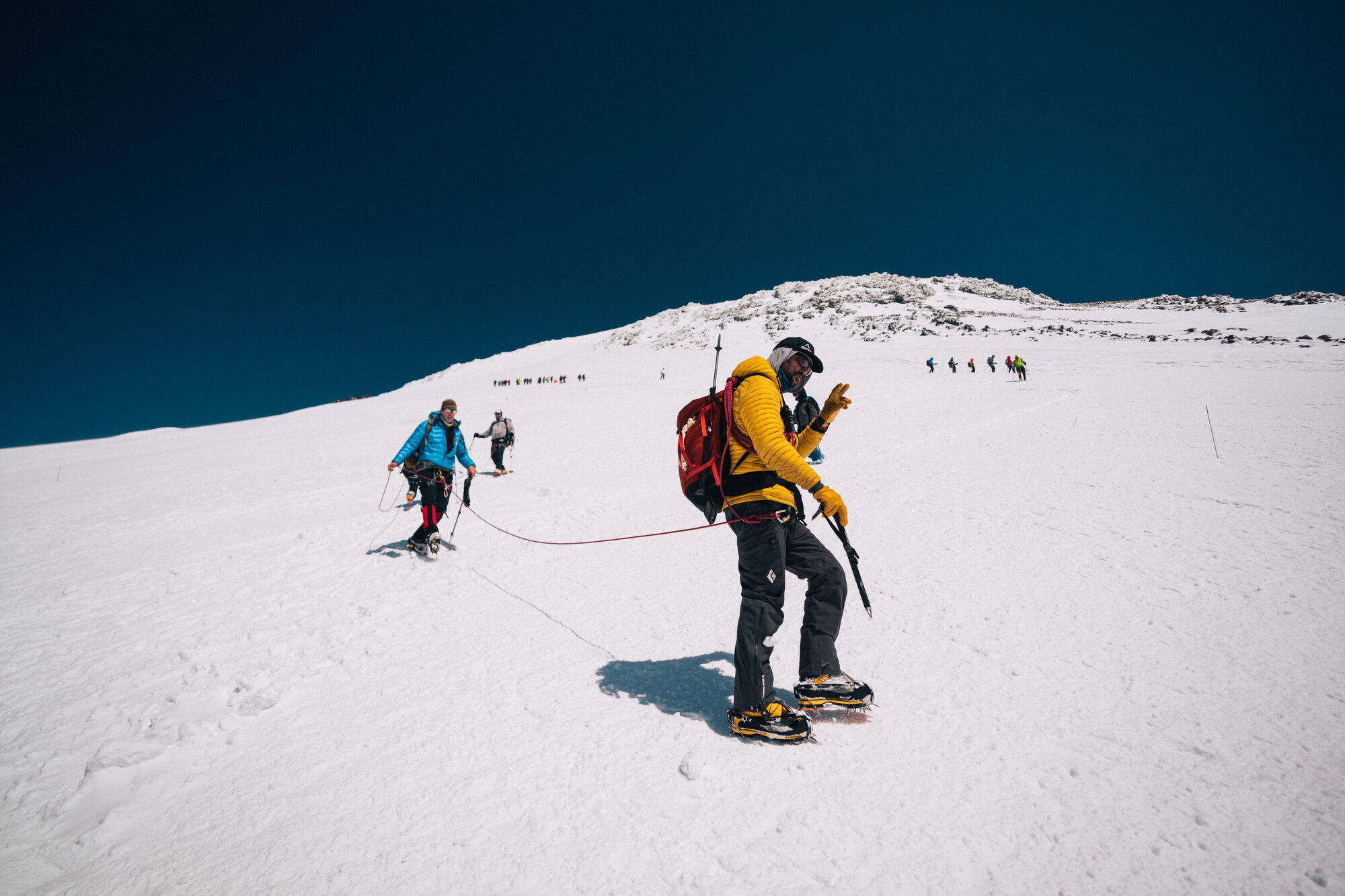 Roped climber in yellow jacket pausing on the snow with crampons biting in and others ahead toward the summit cone