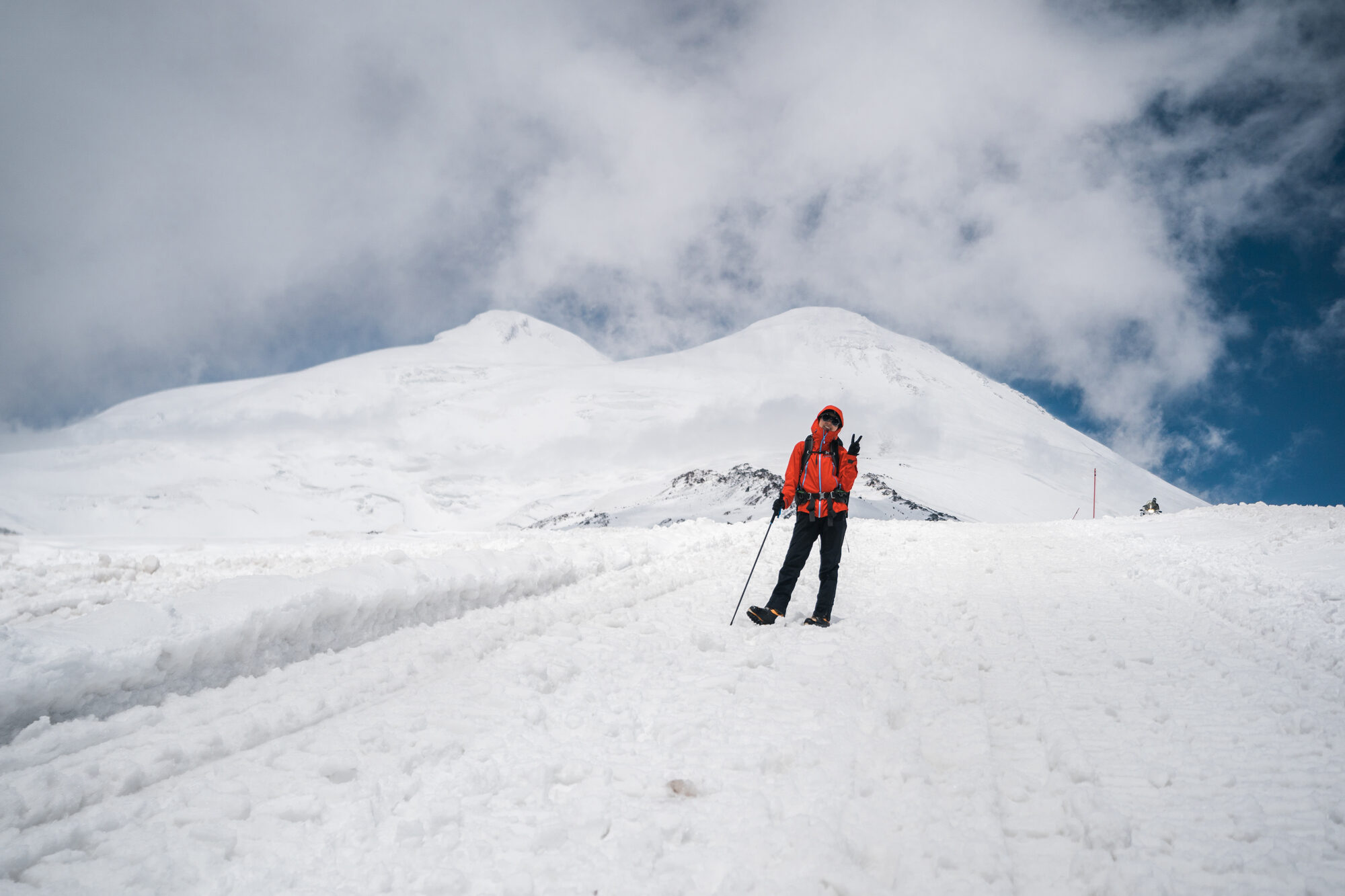 Climber in an orange jacket posing with trekking poles on the snow with both Elbrus summits behind
