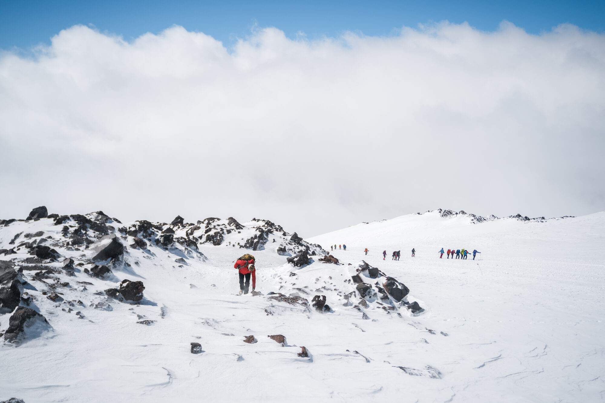 Climber in red passing dark rock outcrops on the snow with a line of climbers in the distance under partly blue sky