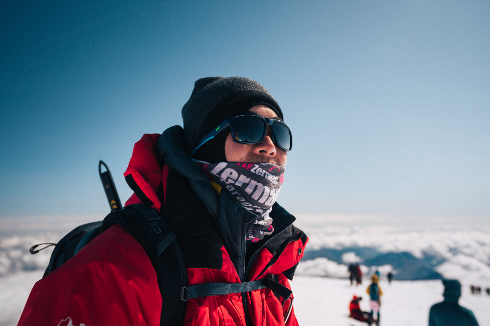 Close portrait of a climber in a red jacket, buff and sunglasses on the summit with other climbers behind