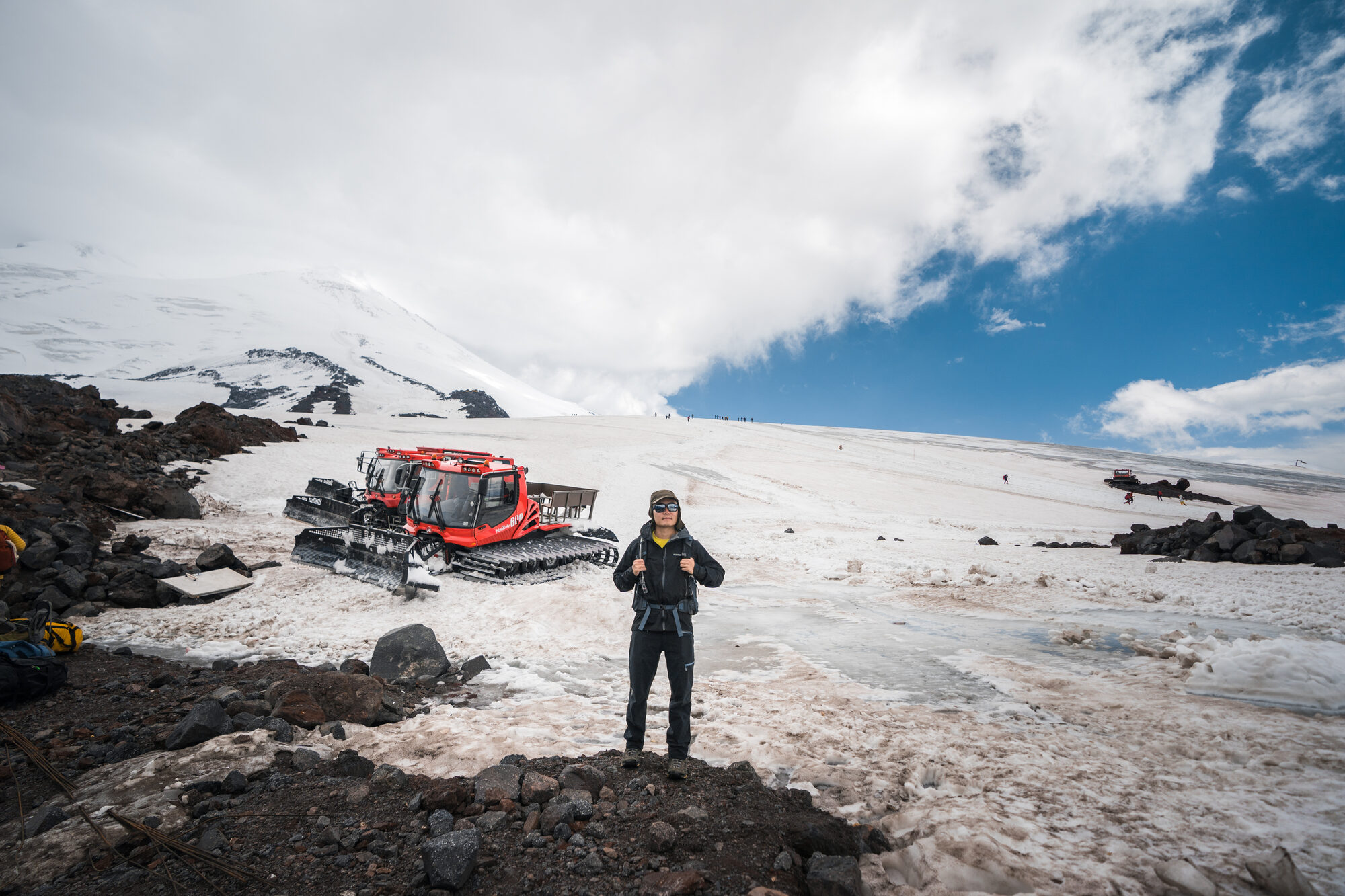 Climber in black kit standing next to one of the red snowcats with the upper mountain and clouds behind