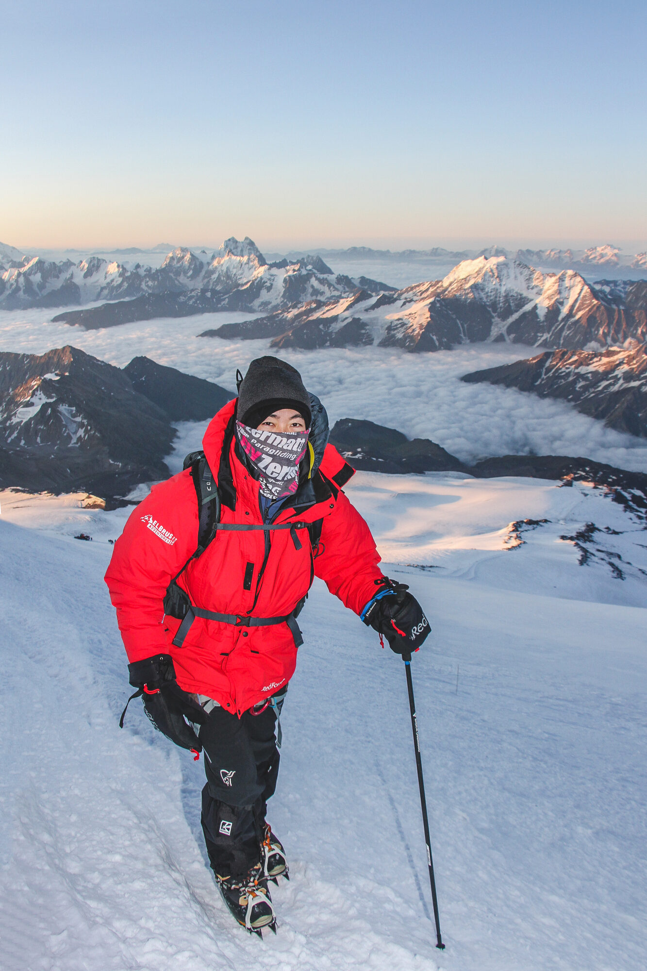 Climber in a red jacket and buff climbing at sunrise with the Caucasus and a sea of cloud behind