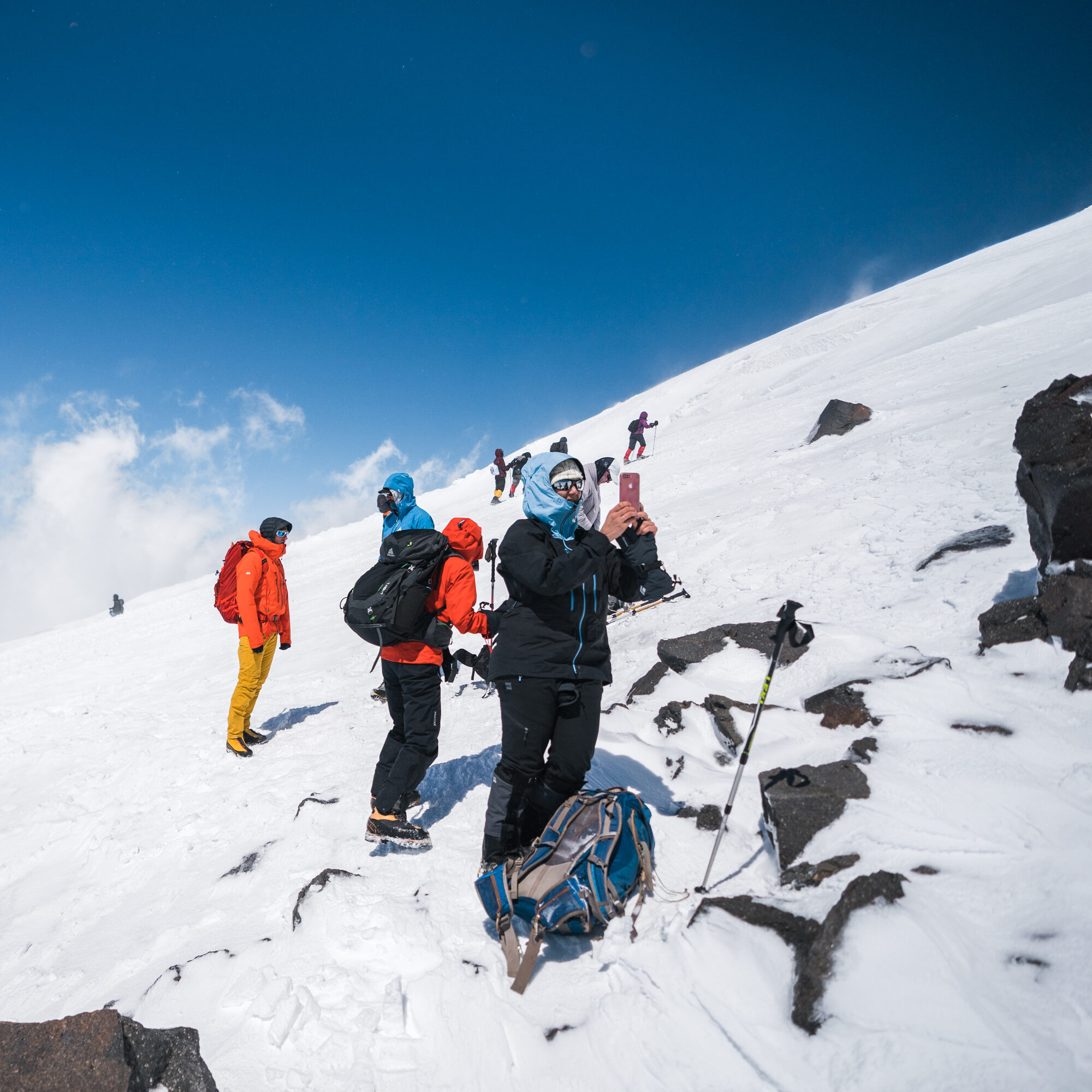 Climbers pausing on a rocky snow slope under deep blue sky, one taking a photo with a phone