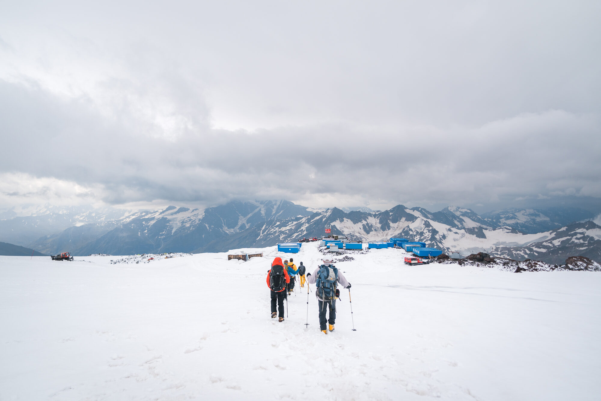 Three climbers walking across the snow toward the base camp containers under grey skies