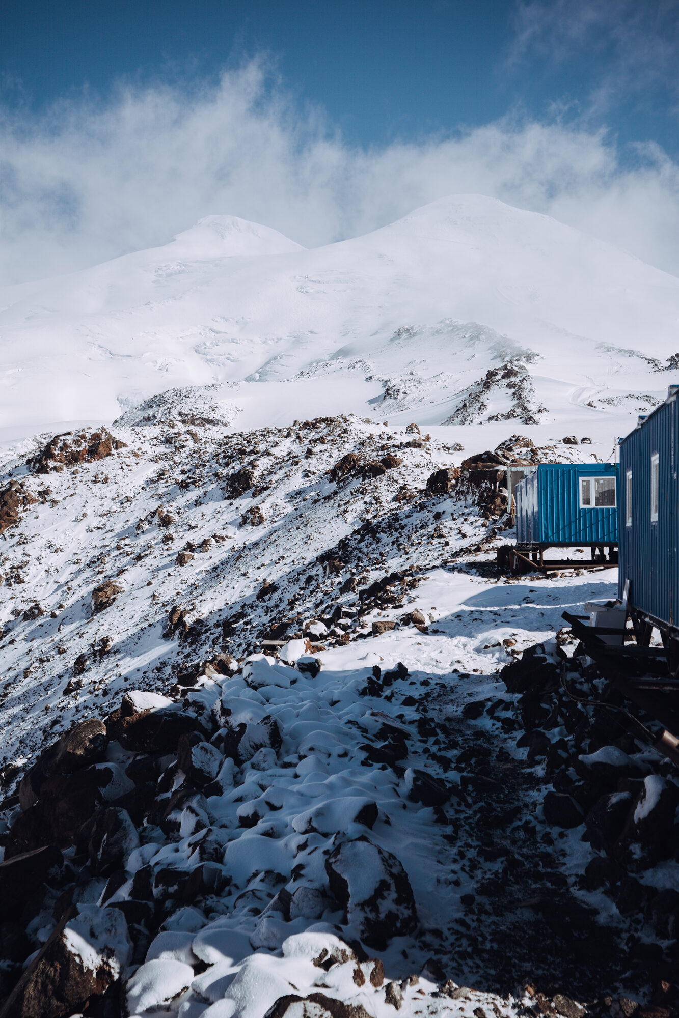 Blue shipping container hut on snowy rocky terrain with Elbrus twin peaks rising behind