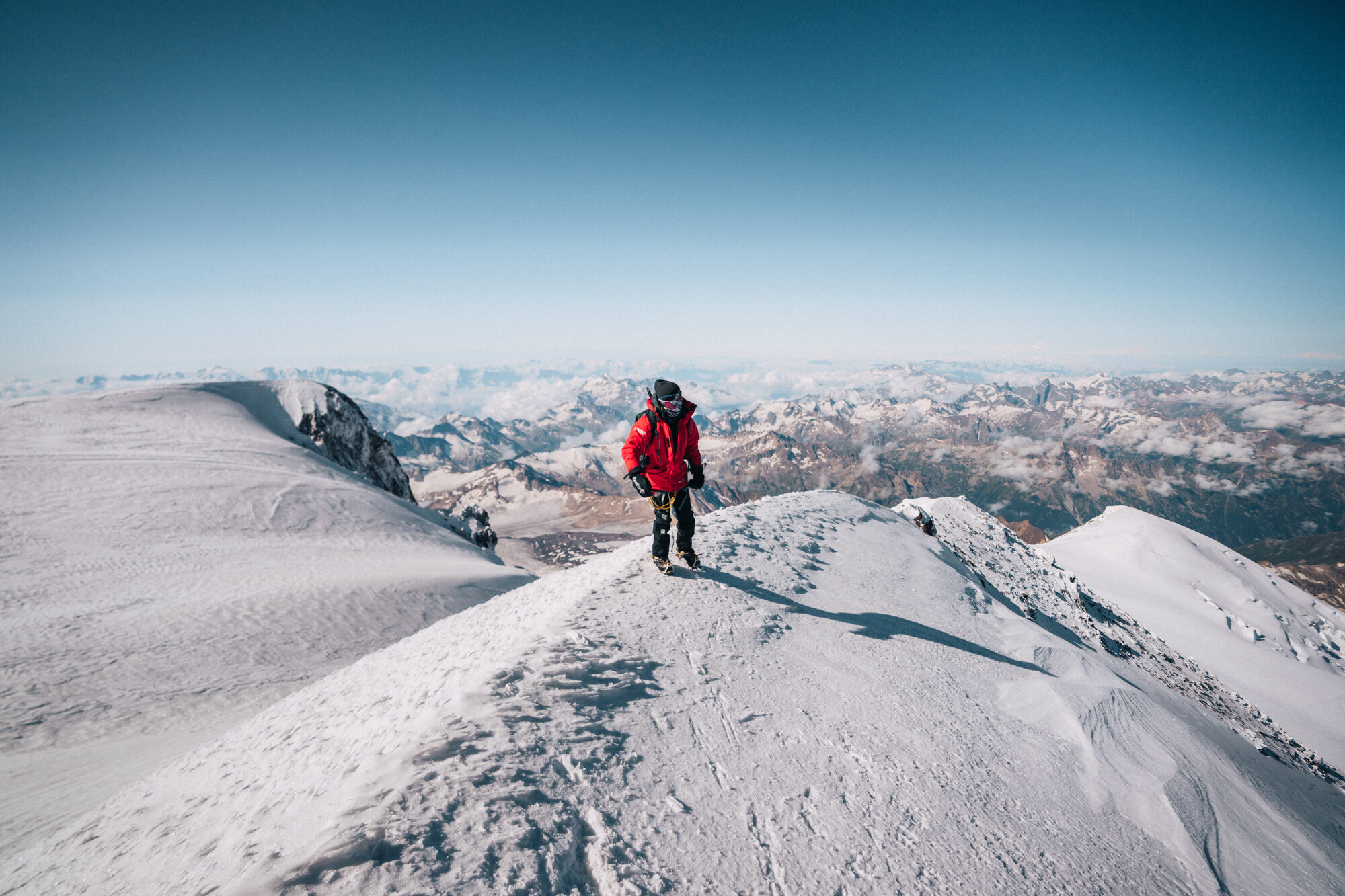 Lone climber crossing the snowy summit ridge of Elbrus with peaks and cloud far below