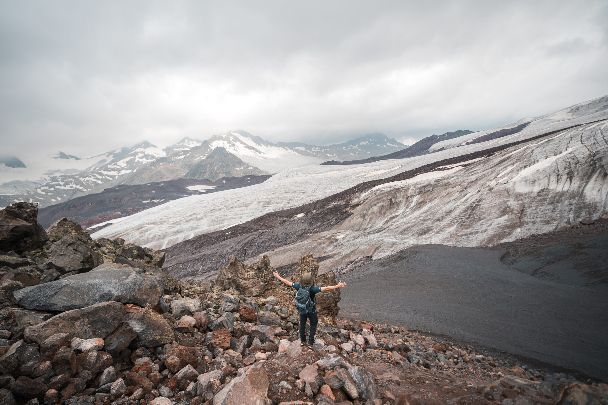 Climber arms outstretched on a rocky moraine with the glacier and snowy peaks spread out behind