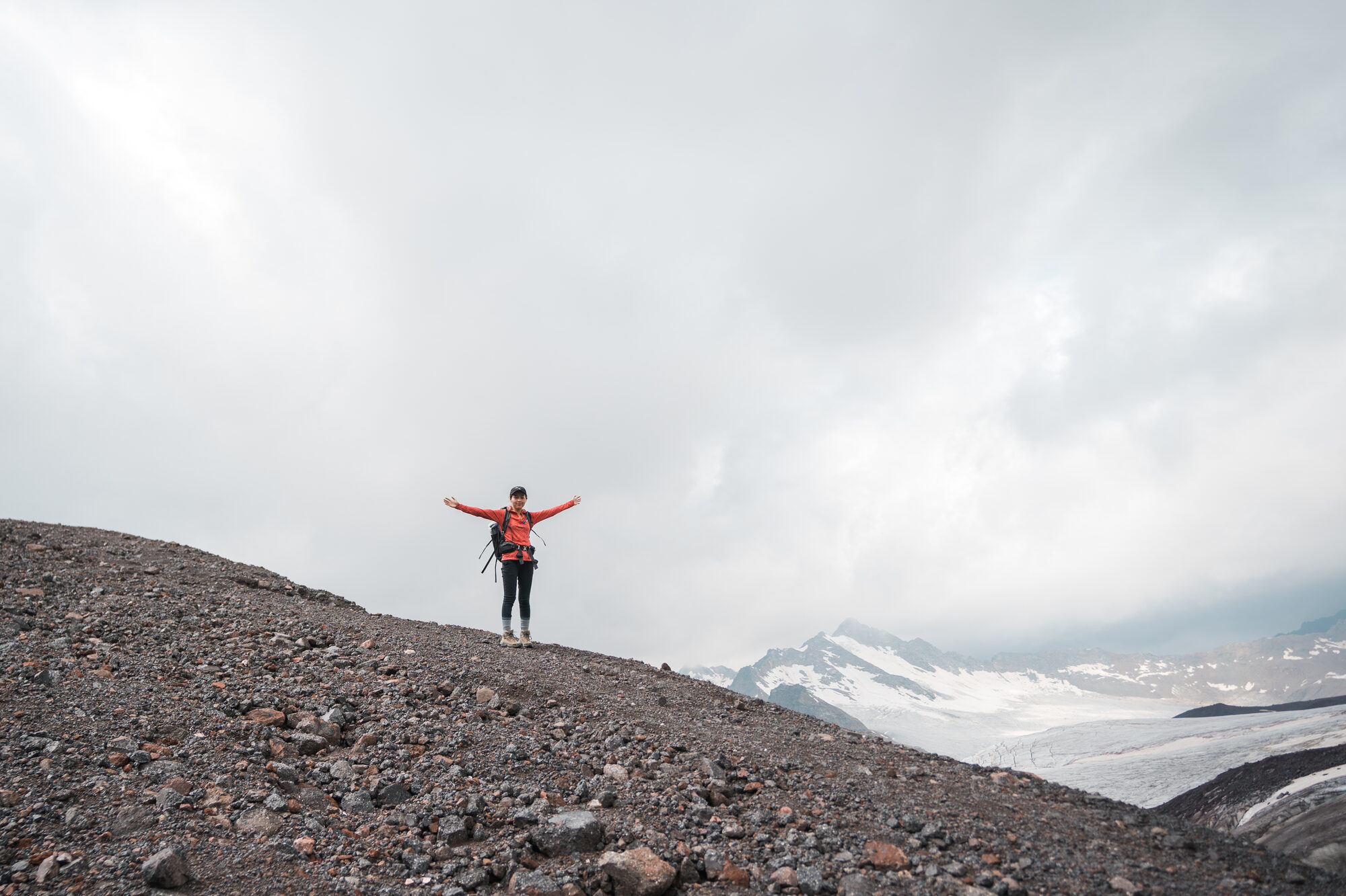 Person with arms outstretched on the gravel slopes of Elbrus with snowy peaks behind