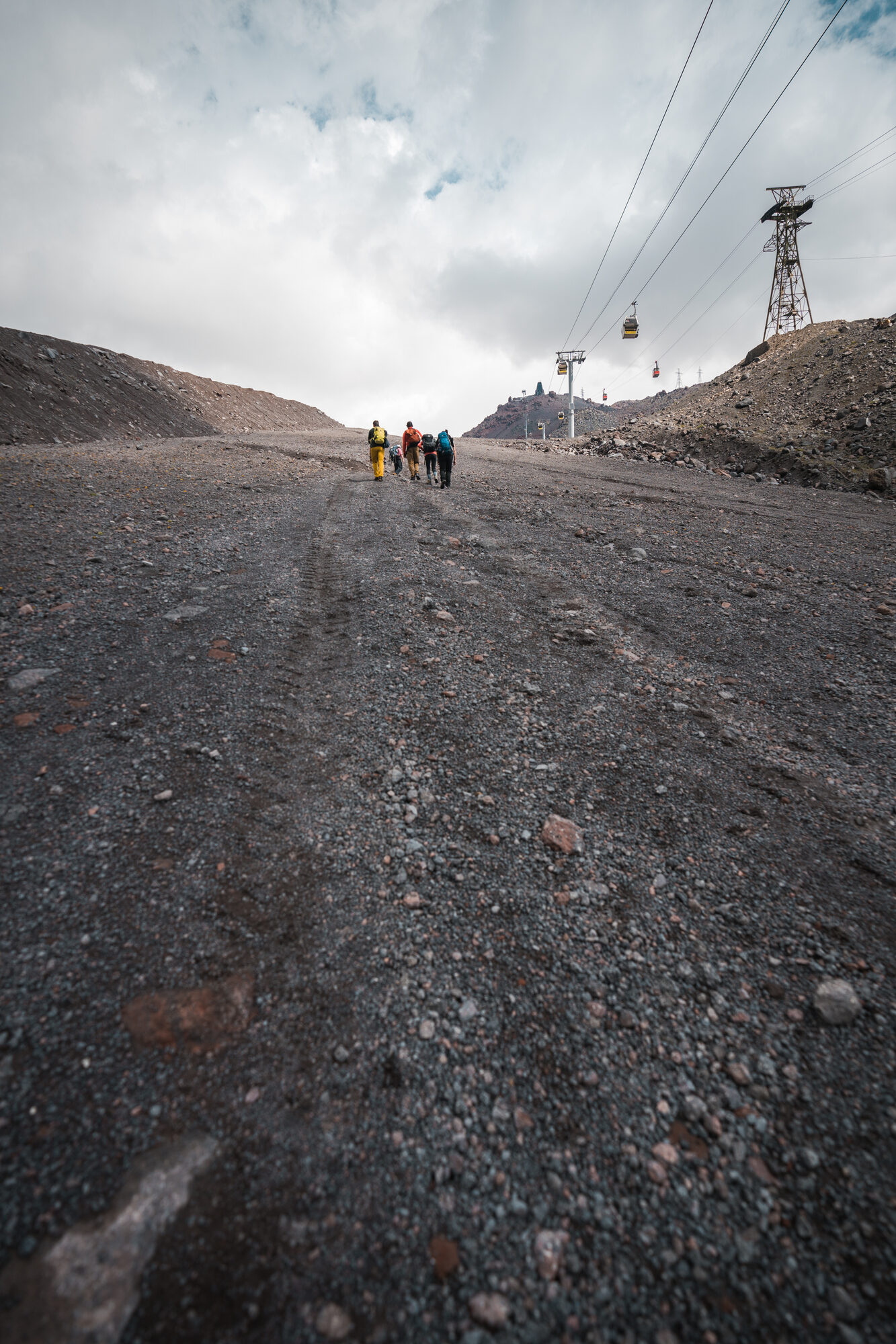 Three climbers walking up a wide gravel track with cable car towers and a passing gondola above