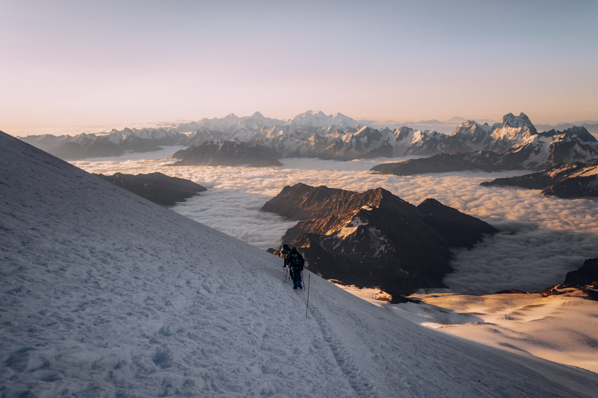Roped pair traversing a steep snow slope at sunrise with the Caucasus glowing gold above a sea of cloud