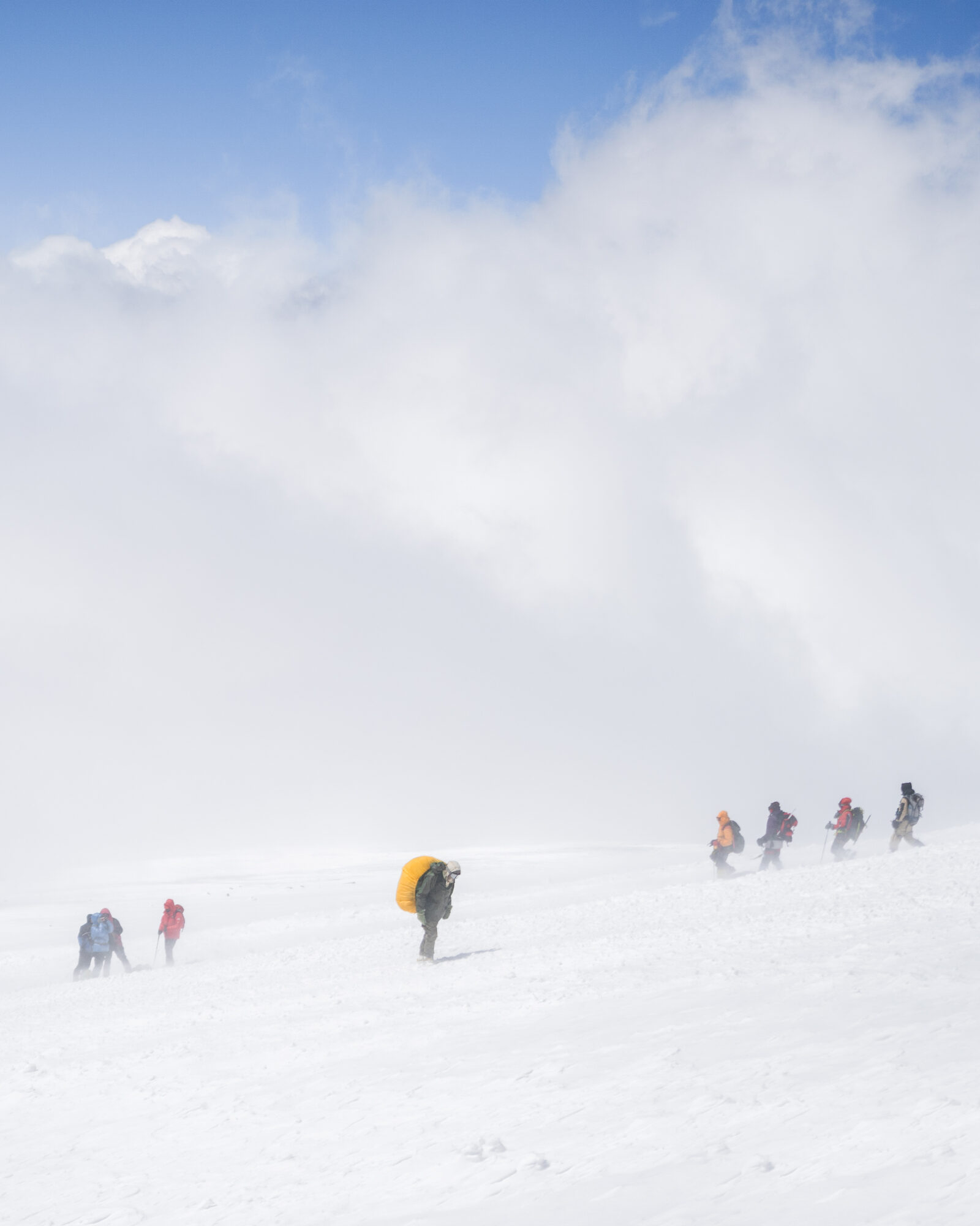 Climbers strung out across a windswept snow slope with cloud streaming over them and only the Elbrus summit cone visible above