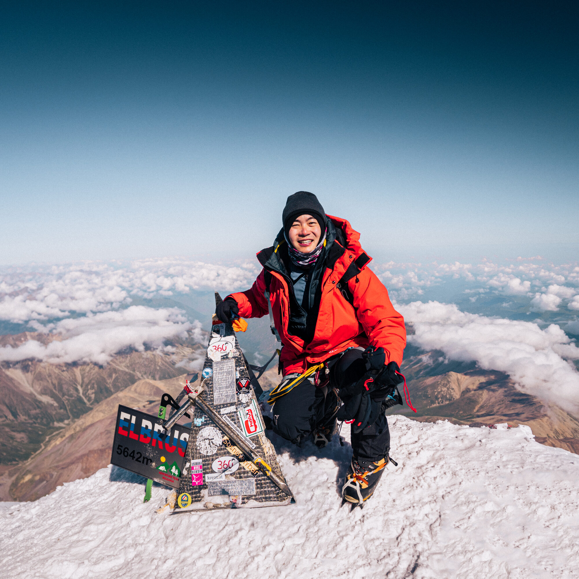 Grace kneeling beside the Elbrus summit sign in a red jacket with clouds and the Caucasus below