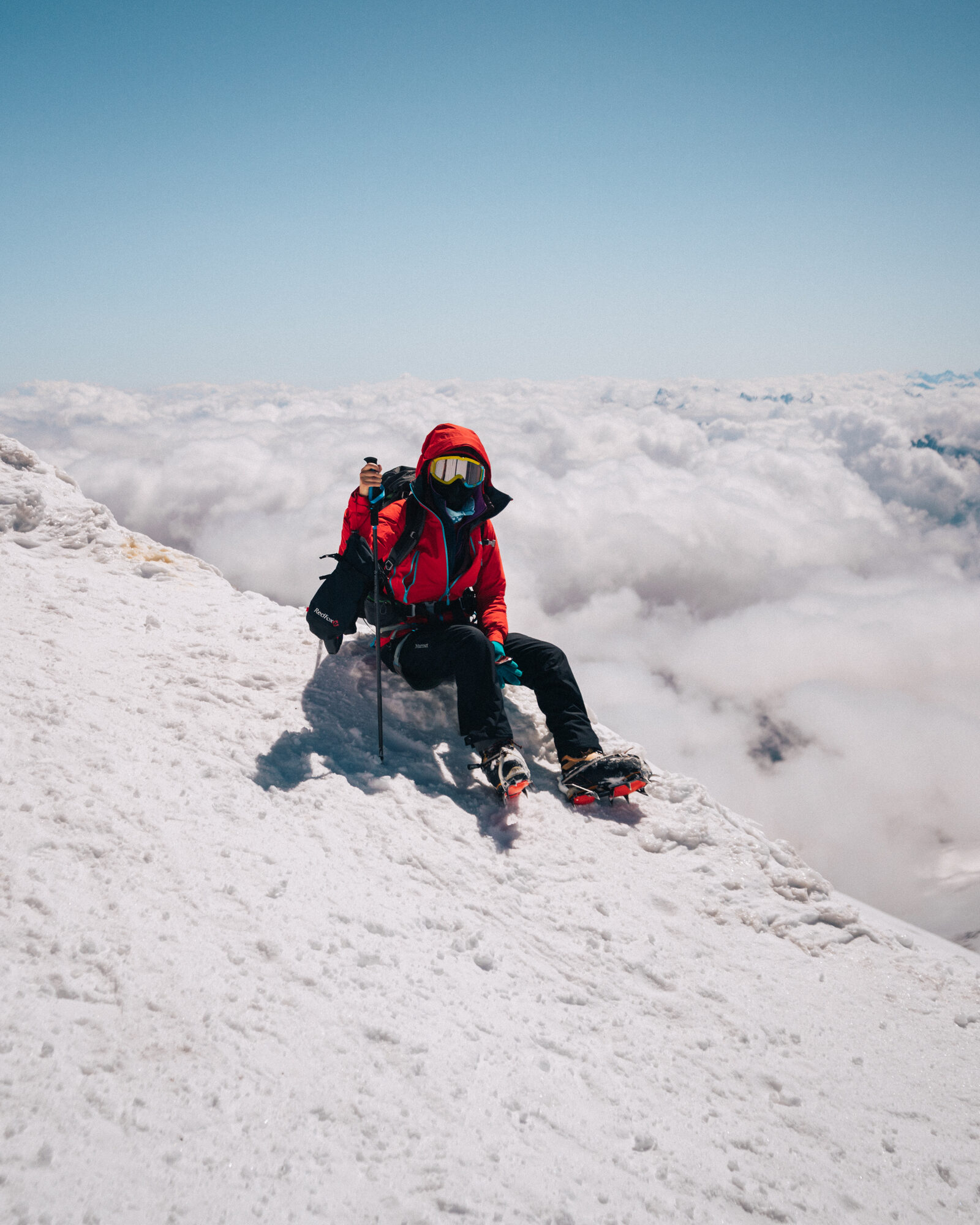 Grace seated on the snow ridge of Elbrus with a sea of clouds and the Caucasus stretching out behind her
