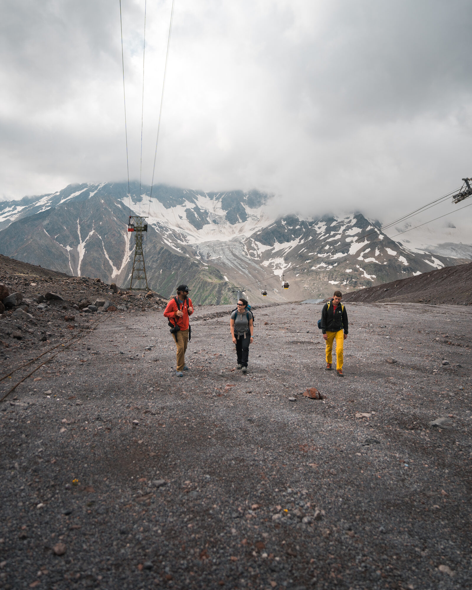 Three climbers walking up the gravel approach with cable car lines and snowy peaks in the clouds