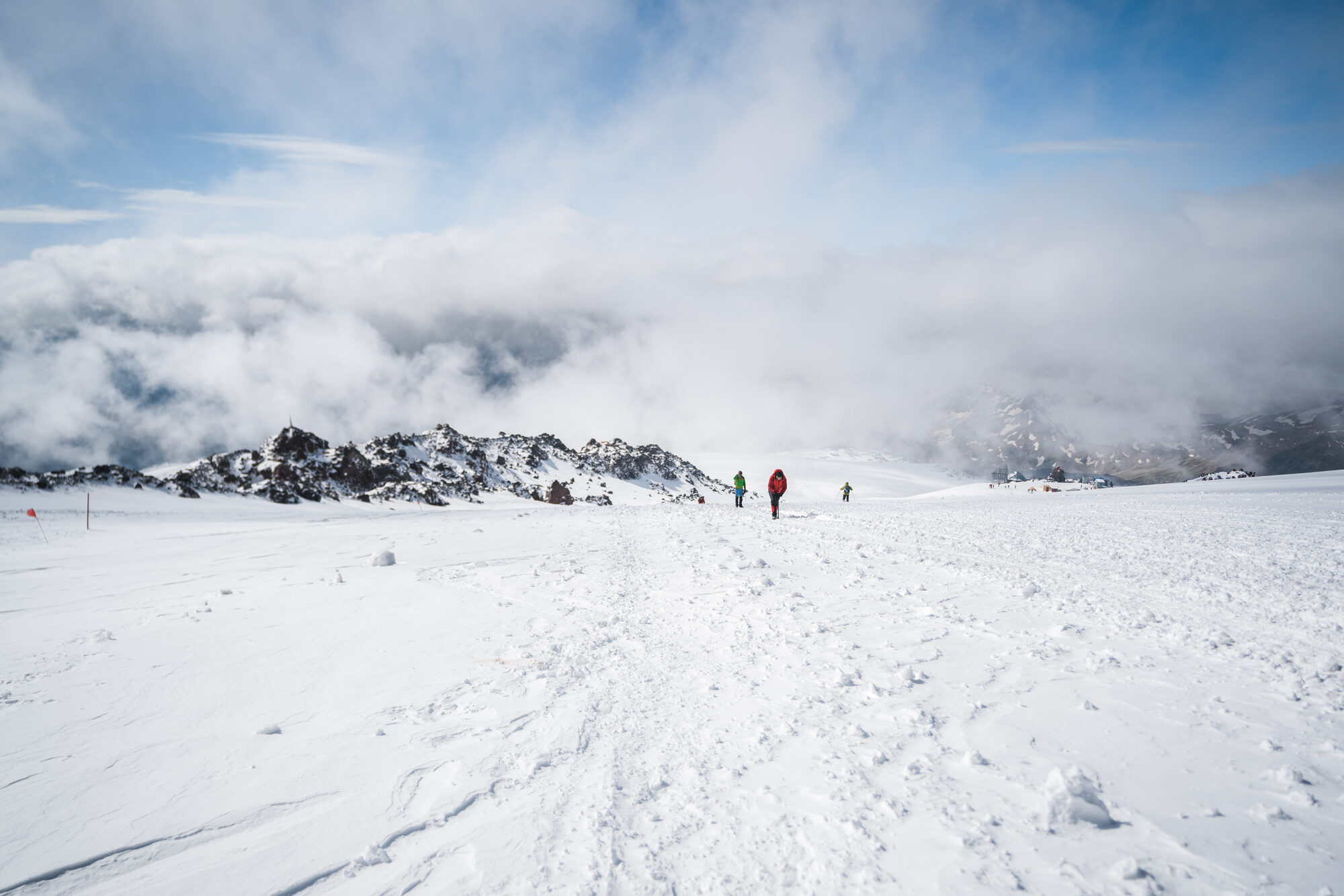 Three climbers on the wide snow plateau next to Pastukhov Rocks with clouds swirling below the peaks