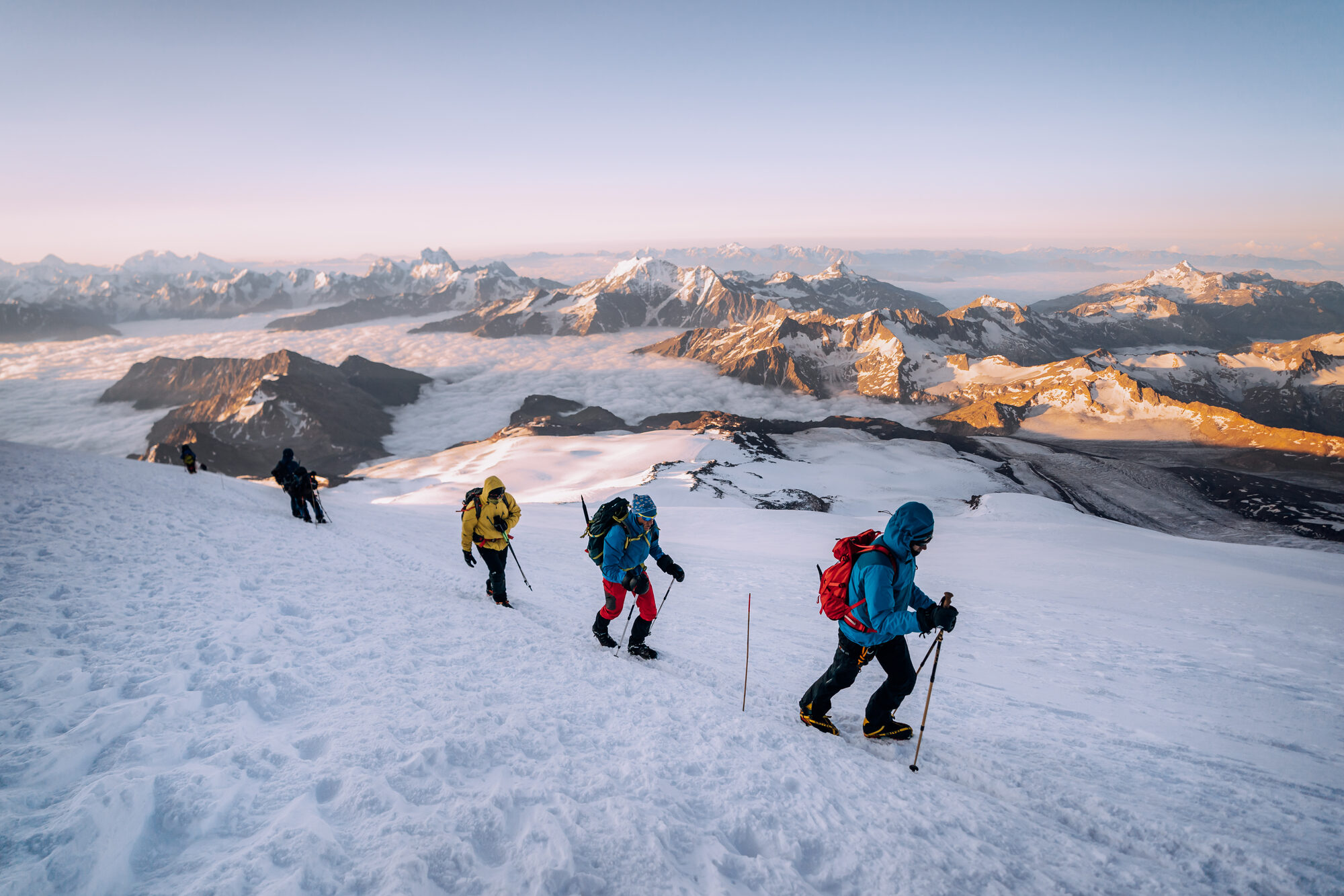 Climbers in single file traversing the snow slope at sunrise with the Caucasus lit gold behind