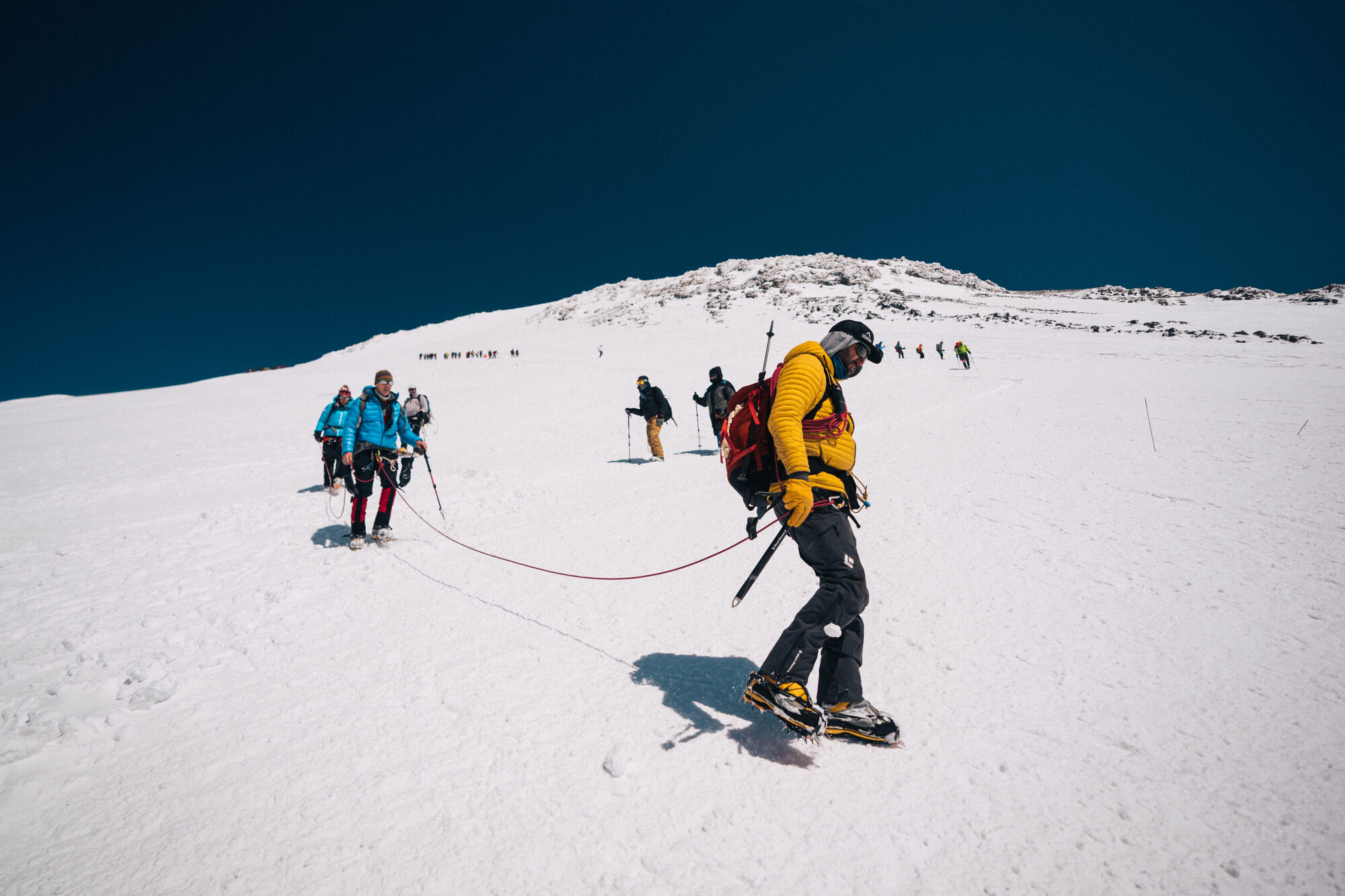 Roped climber in a yellow jacket crossing the summit slope with teammates on the rope behind