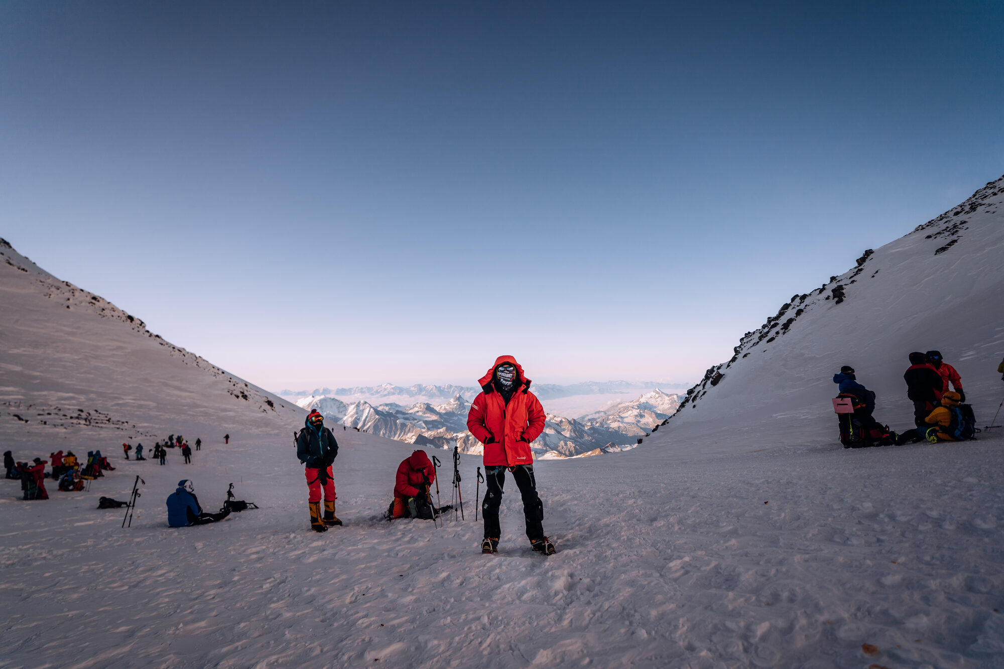 Climber in red jacket pausing in the saddle between Elbrus's twin peaks with other climbers resting around