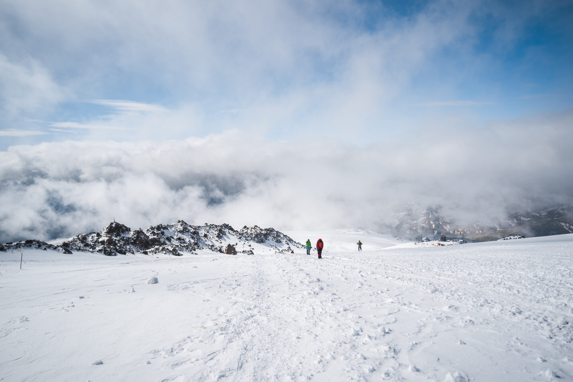 Small group of climbers on the snow plateau beside dark rock outcrops with cloud banked below