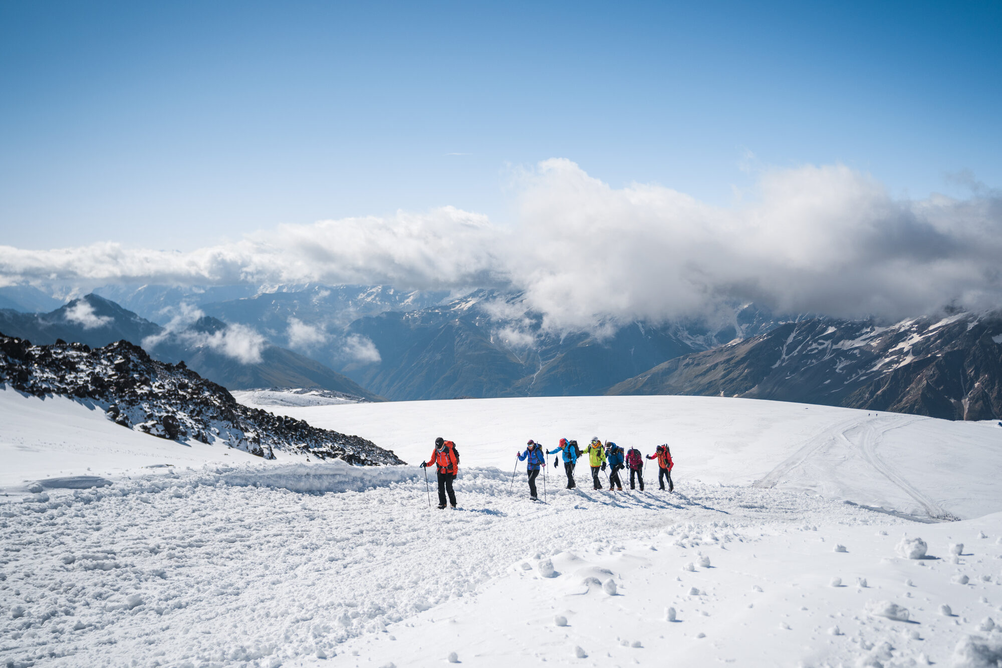 Team in single file across a wide snow plateau with clouds banked below and peaks in the distance