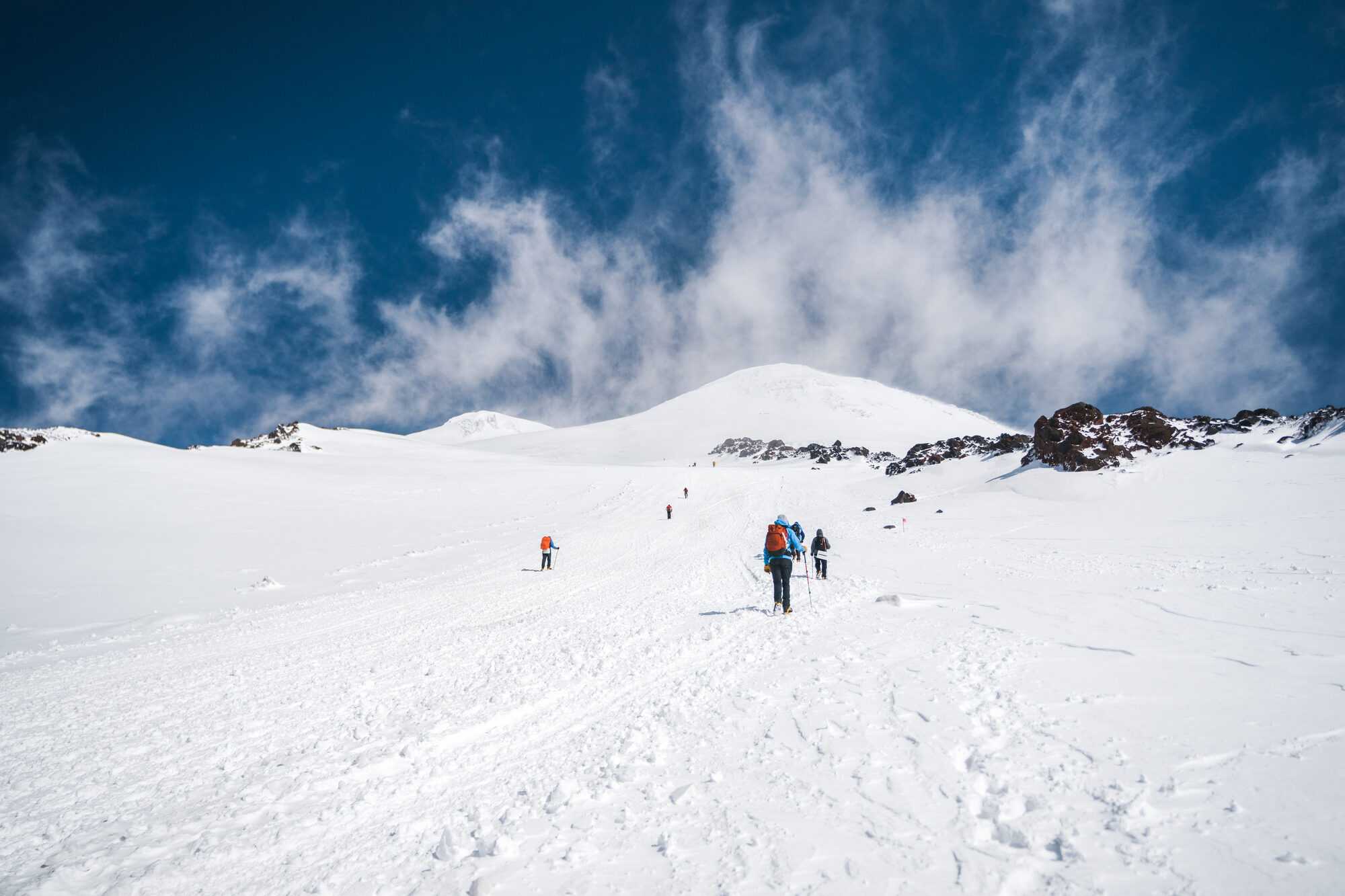 Climbers spread out on a snow slope under blue sky with the Elbrus summit cone behind
