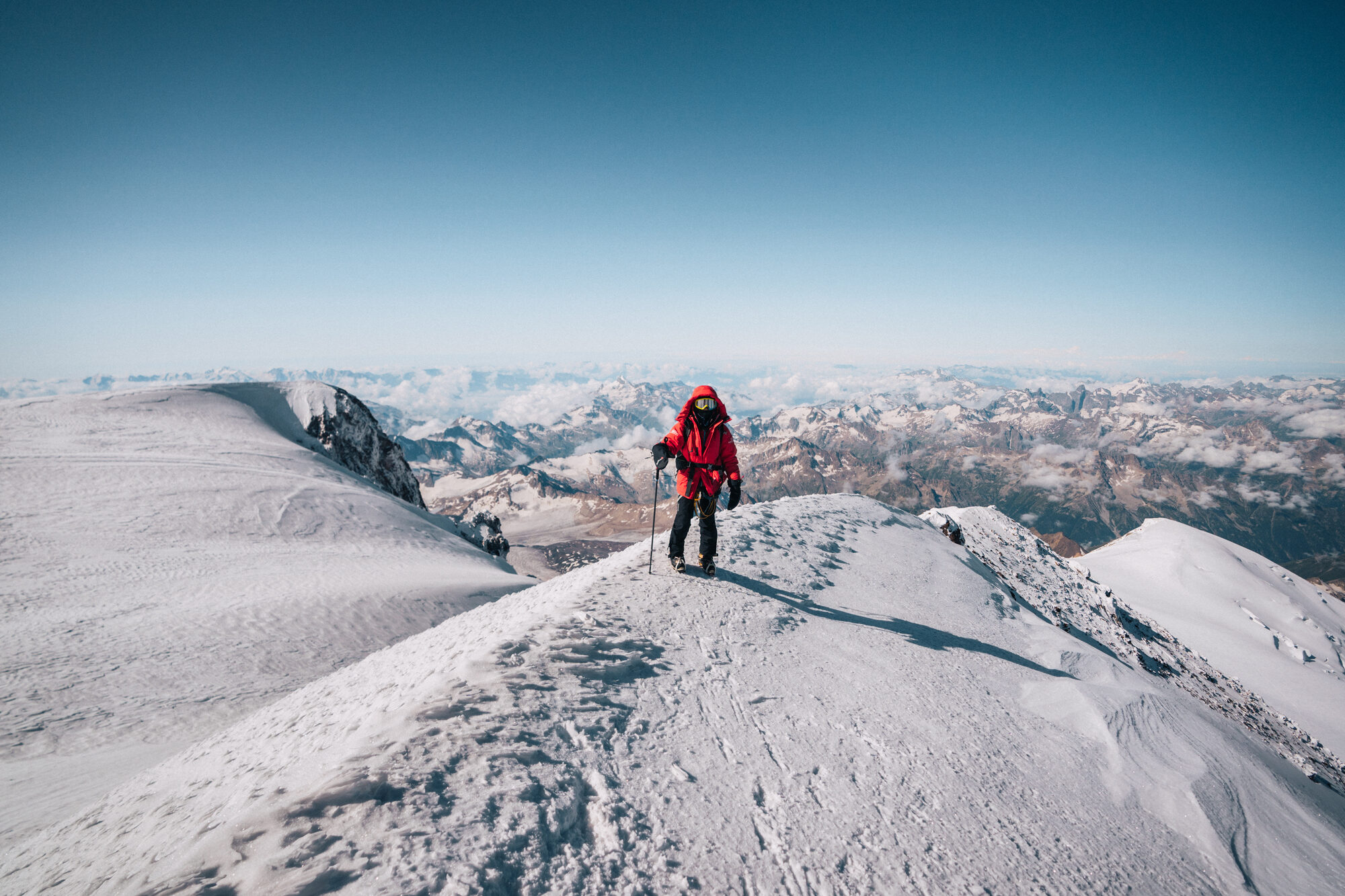 Climber in red descending the snowy summit ridge with the Caucasus spread out behind