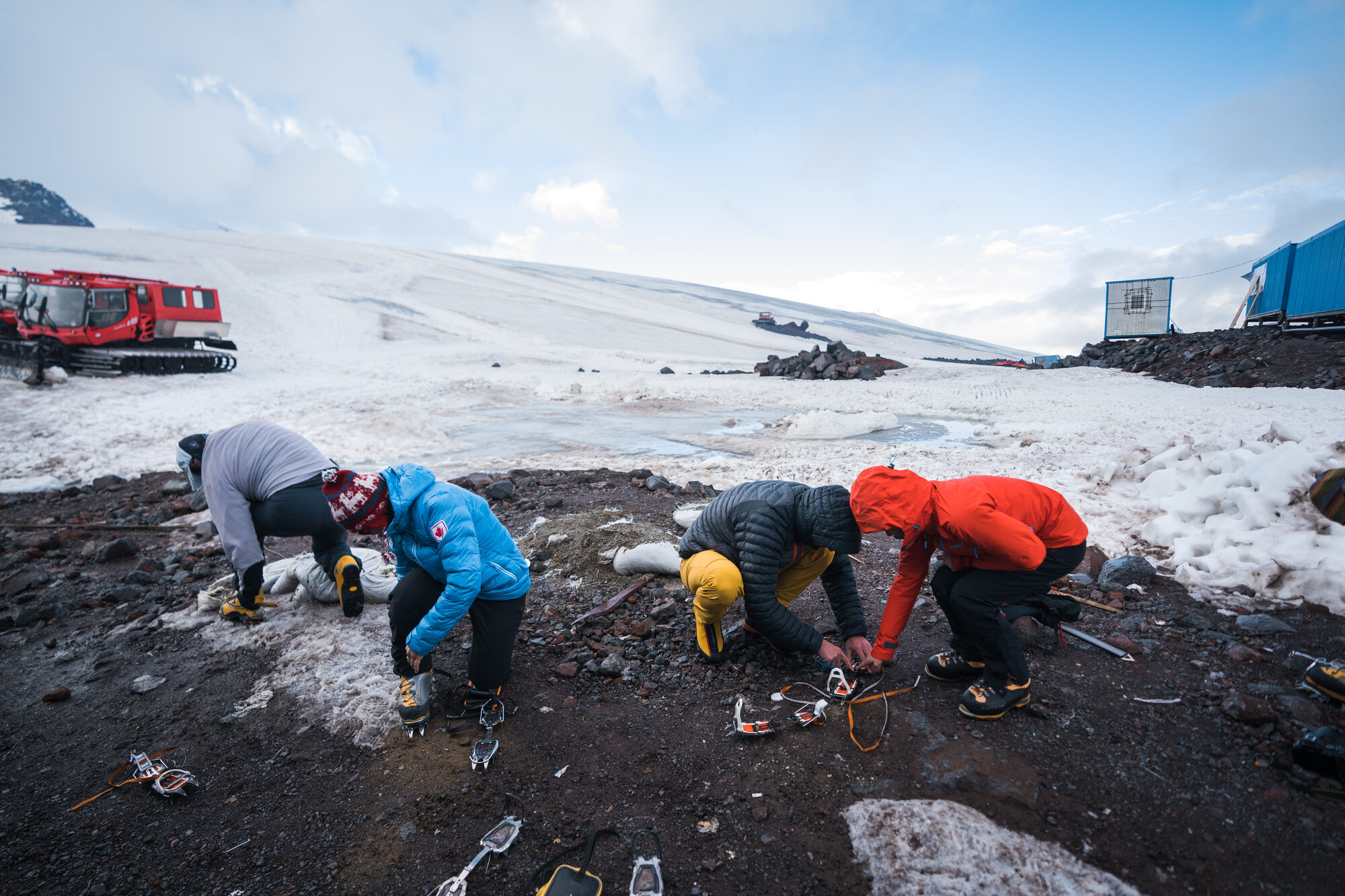 Team bent over on the rocky snow near base camp strapping crampons onto their boots