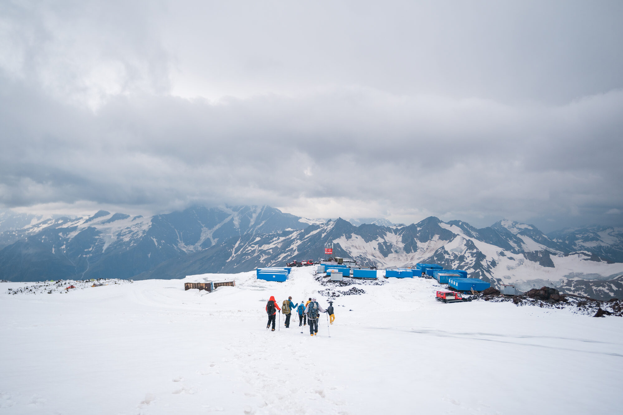 Team hiking through snow toward Elbrus base camp with containers visible