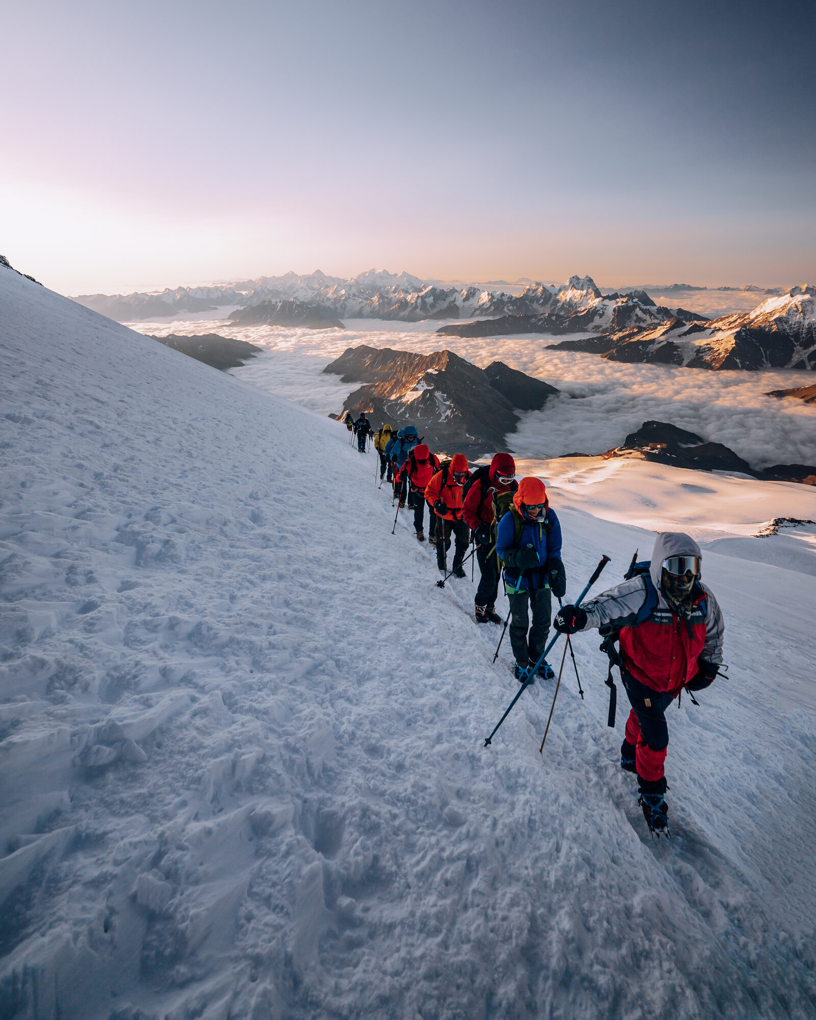 Line of climbers in single file ascending a snow slope at sunrise with the Caucasus lit gold beyond