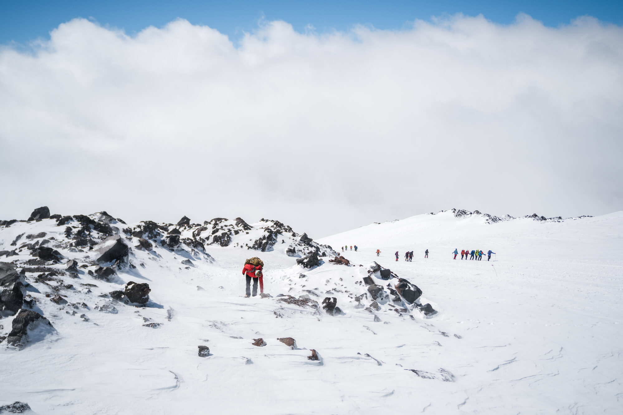 Team in a long line returning across a wide snow field with dark rock outcrops on the left