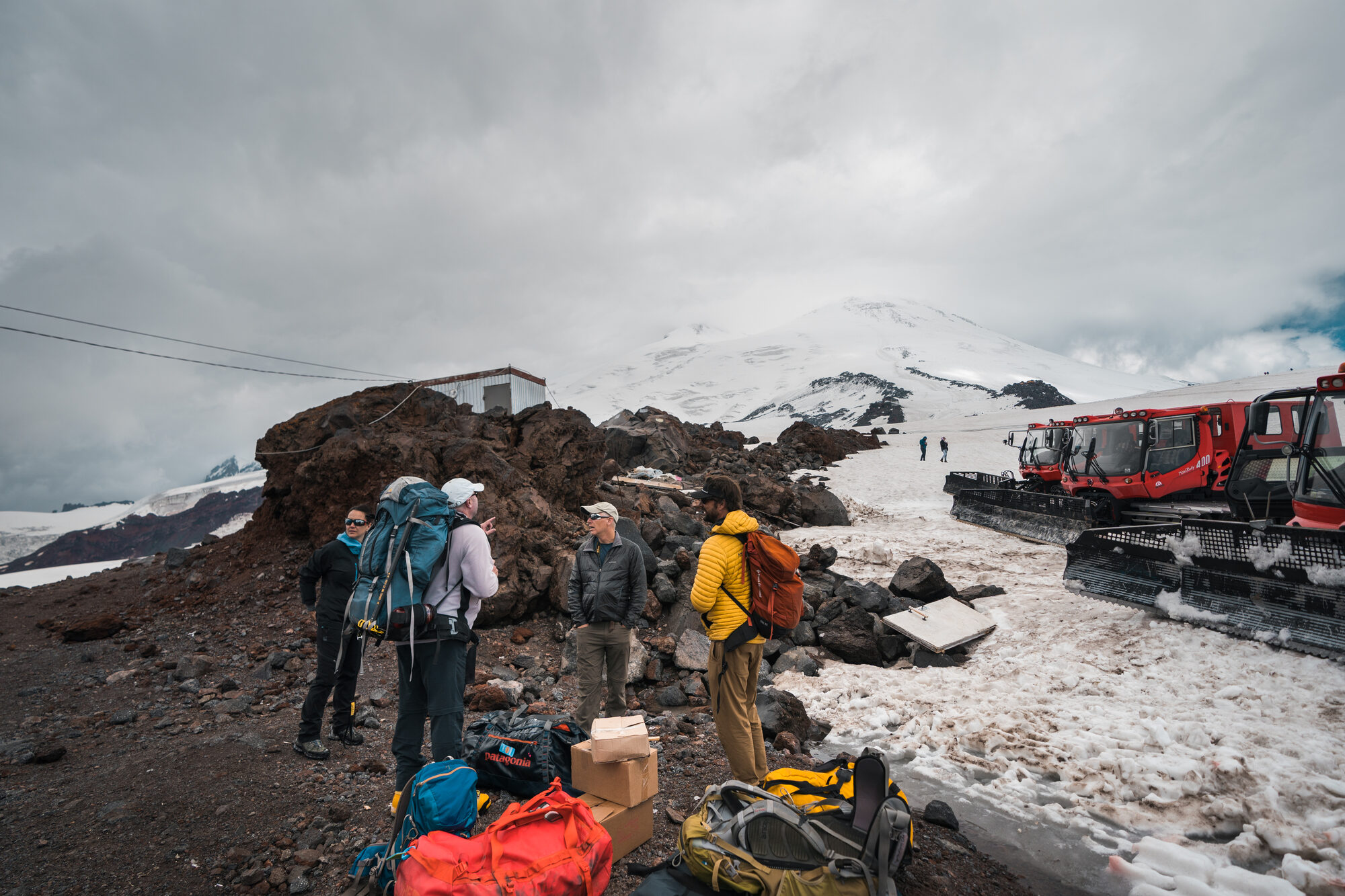 Team sorting duffle bags on the rocks at base camp with red snowcats parked behind