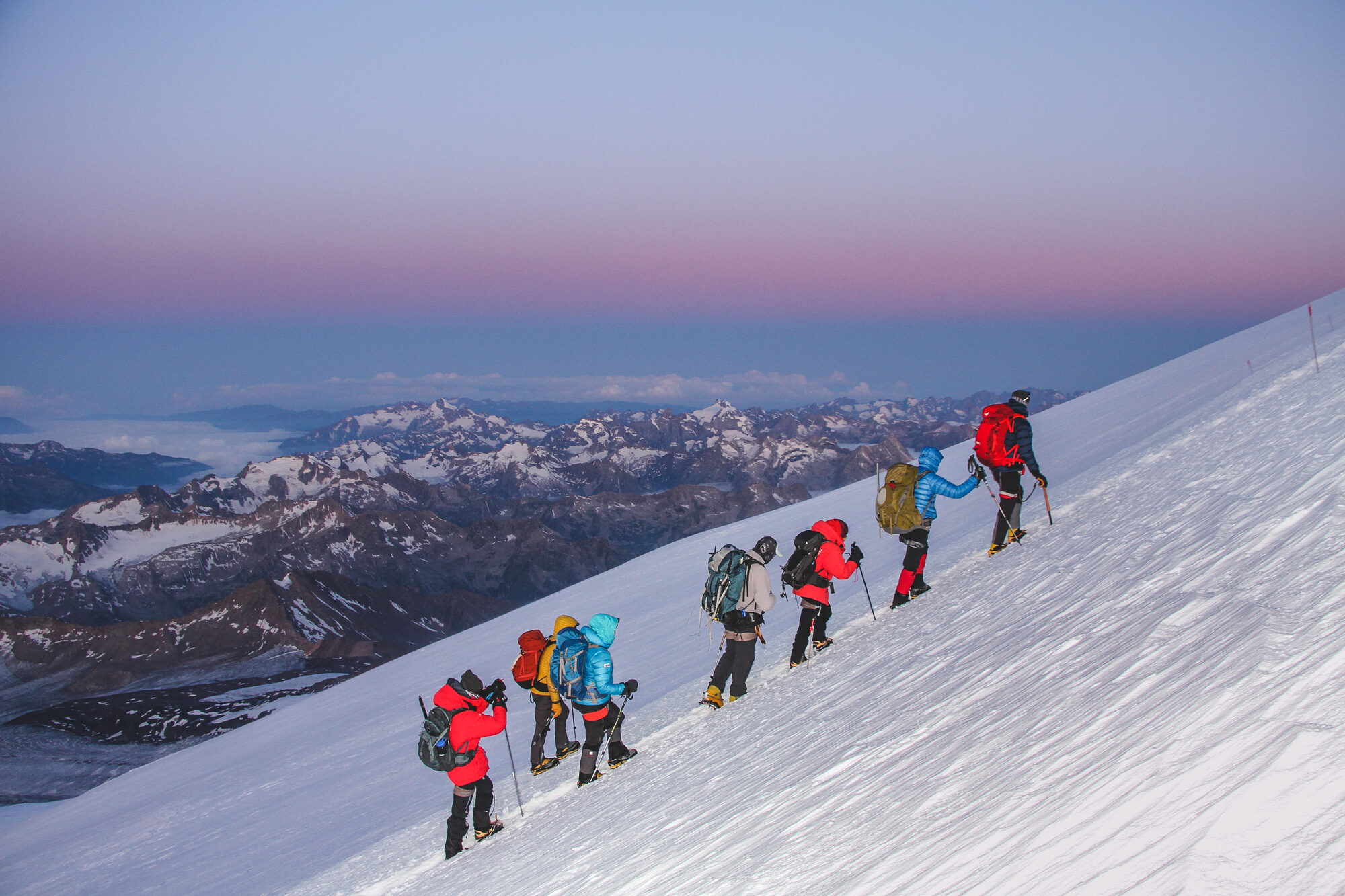 Six climbers in single file ascending the snow slope at dawn with pink light over the Caucasus