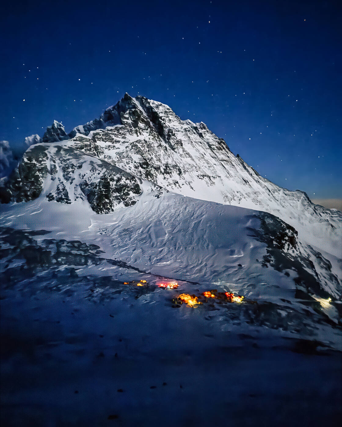 Night aerial drone shot of a snow-covered mountain peak illuminated by camp lights below with stars above