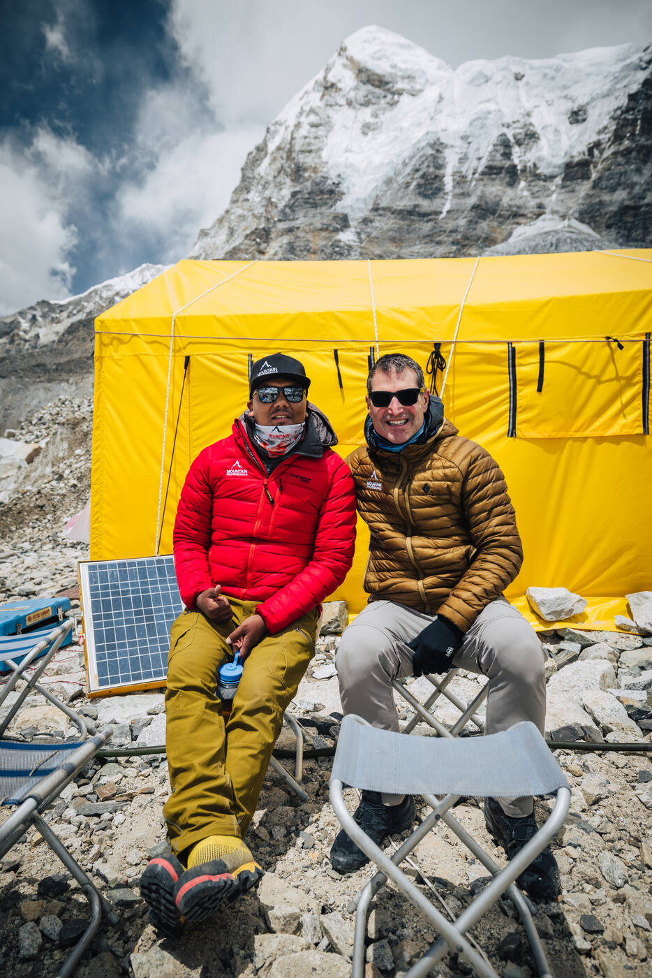 Two team members sitting together at Everest Base Camp with solar panel and yellow tent behind
