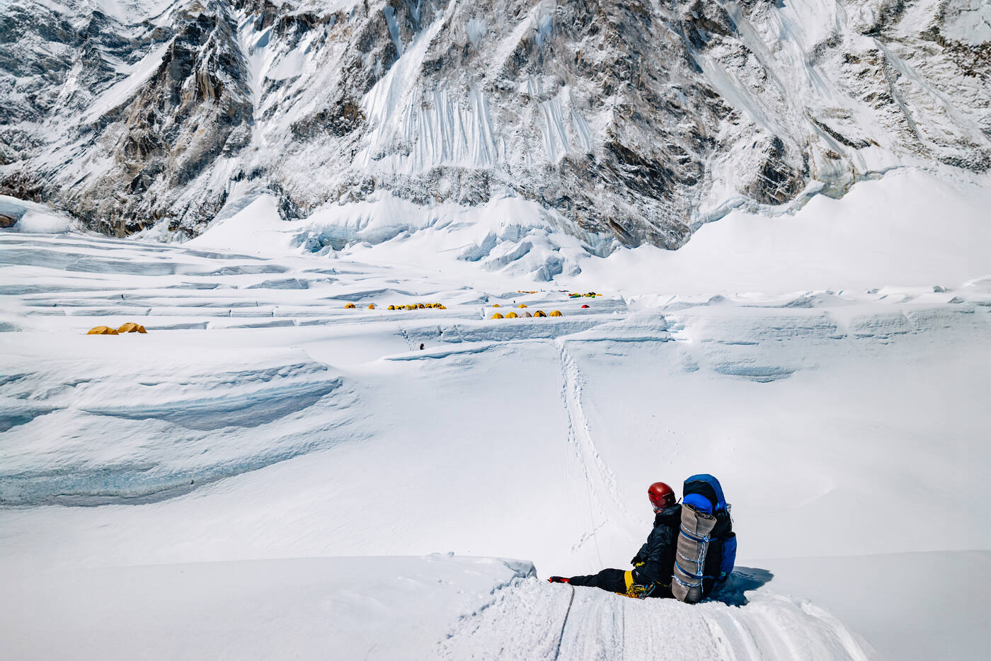 A climber sitting on the snow with tents of a high camp and mountain walls behind
