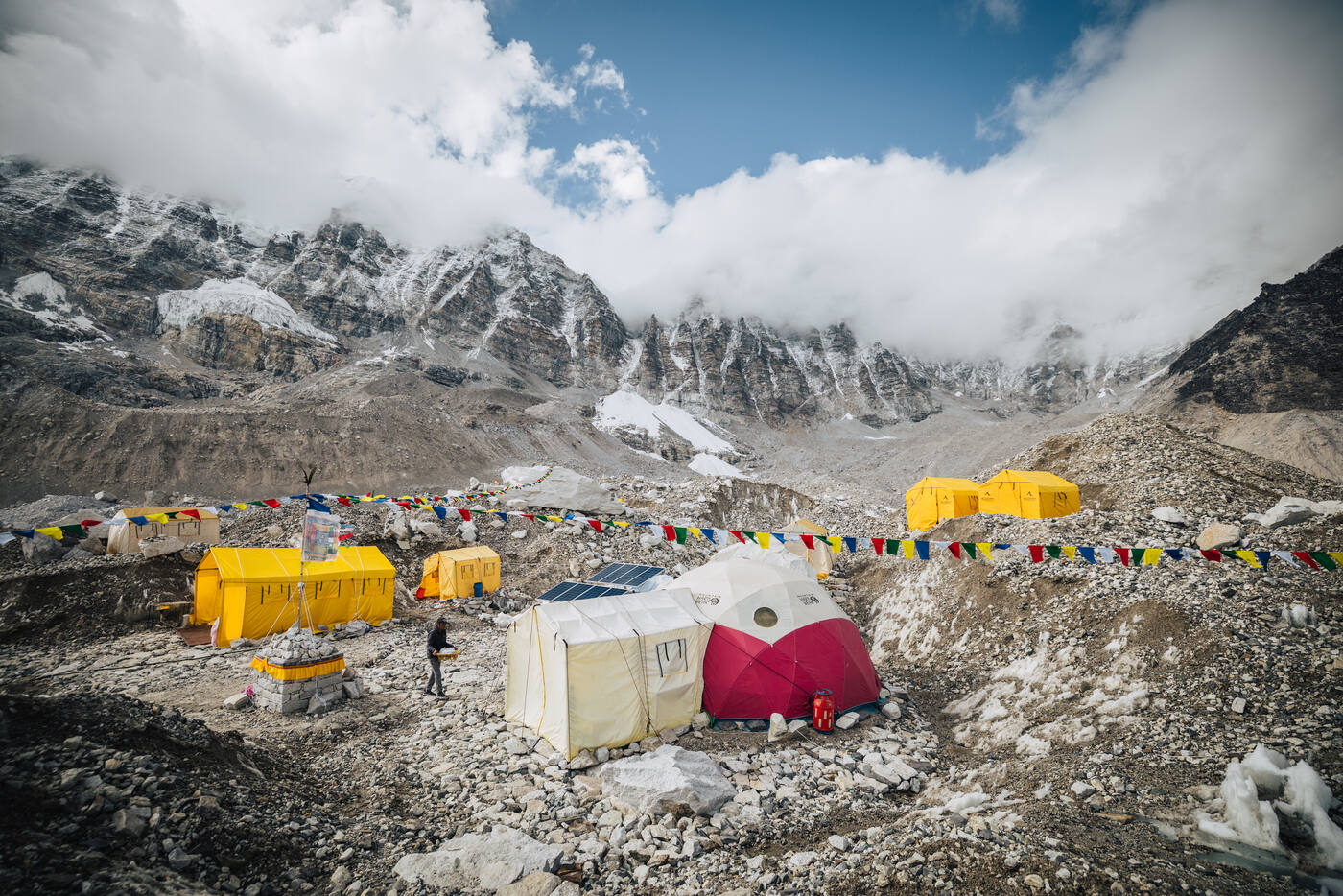 Mountain Professionals camp with yellow tents, prayer flags, and a solar panel beneath snow-covered peaks