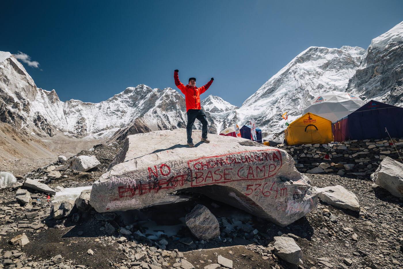 A climber in a red jacket standing with arms raised on the Everest Base Camp rock, tents and peaks behind