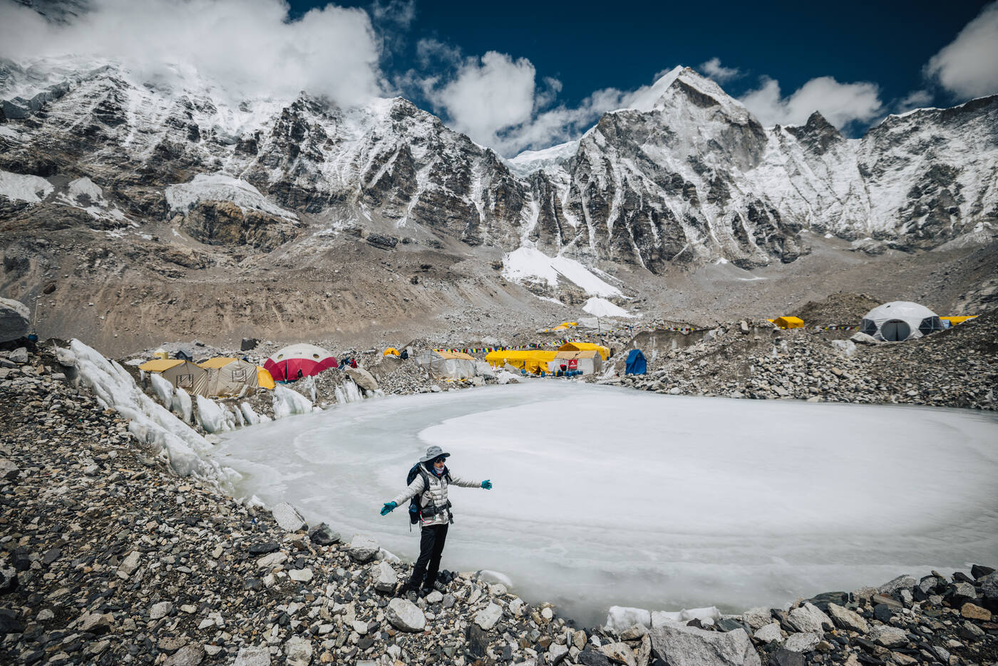 A climber standing beside a frozen glacial pond at Everest Base Camp with tents and peaks behind