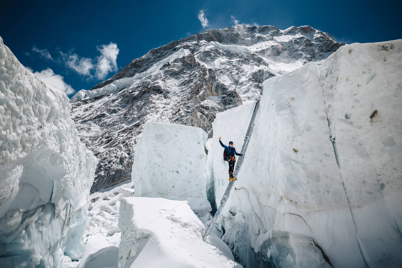 A climber waving from a ladder propped against an ice block in the Khumbu Icefall
