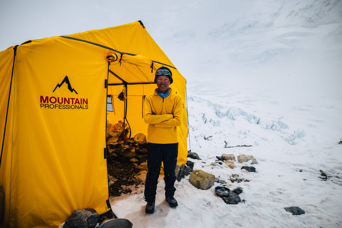 A climber in a yellow jacket standing outside a yellow Mountain Professionals tent on snow