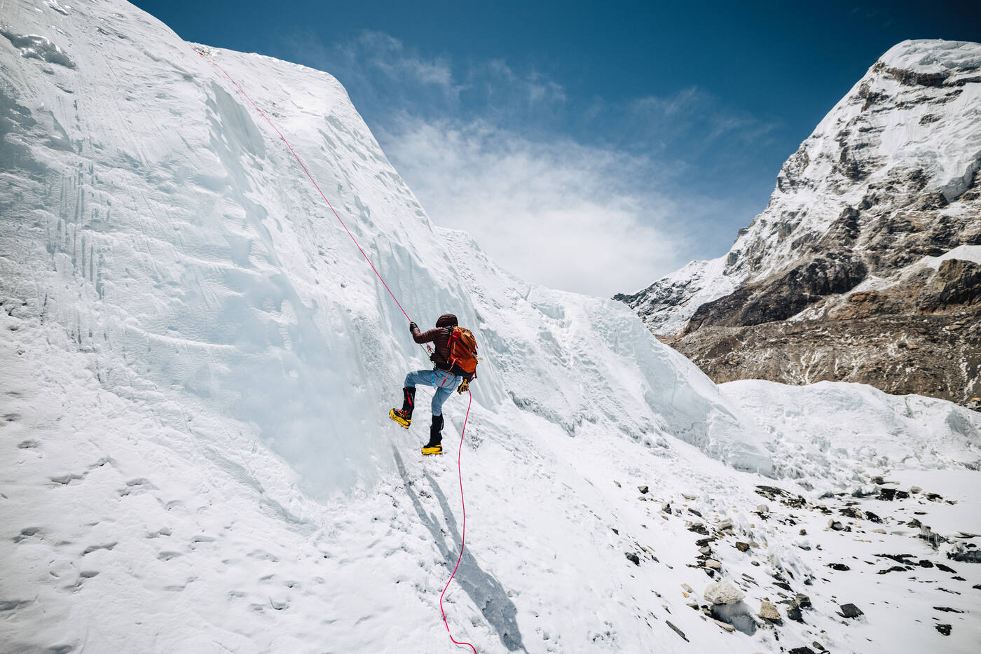 A climber ascending a steep ice wall on a fixed rope with mountains behind
