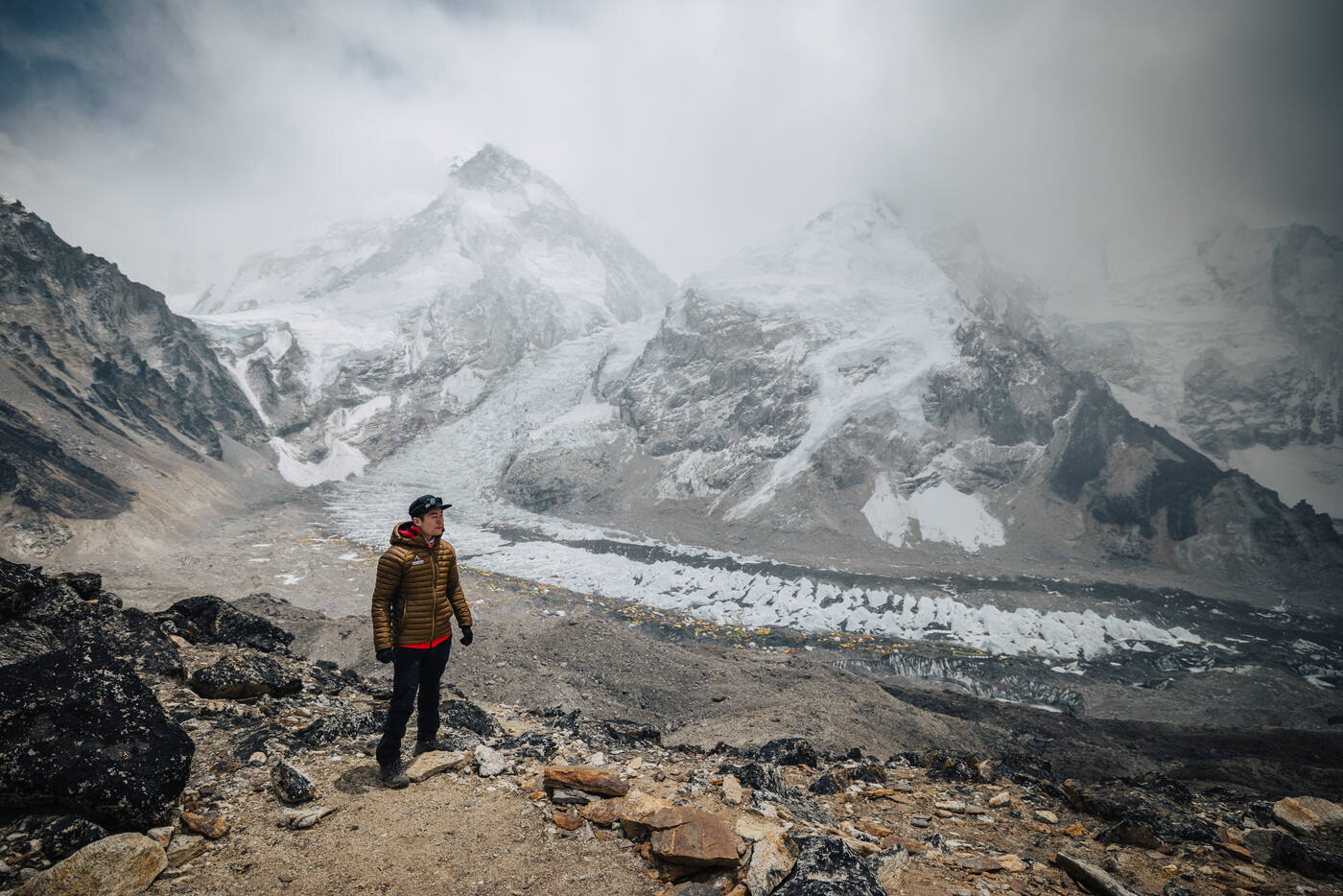 A climber in a yellow jacket standing on a rocky hillside above Base Camp, looking toward the Khumbu Icefall