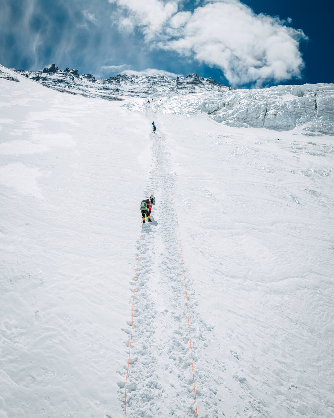 Climbers ascending a fixed line up a broad snow slope beneath ice cliffs