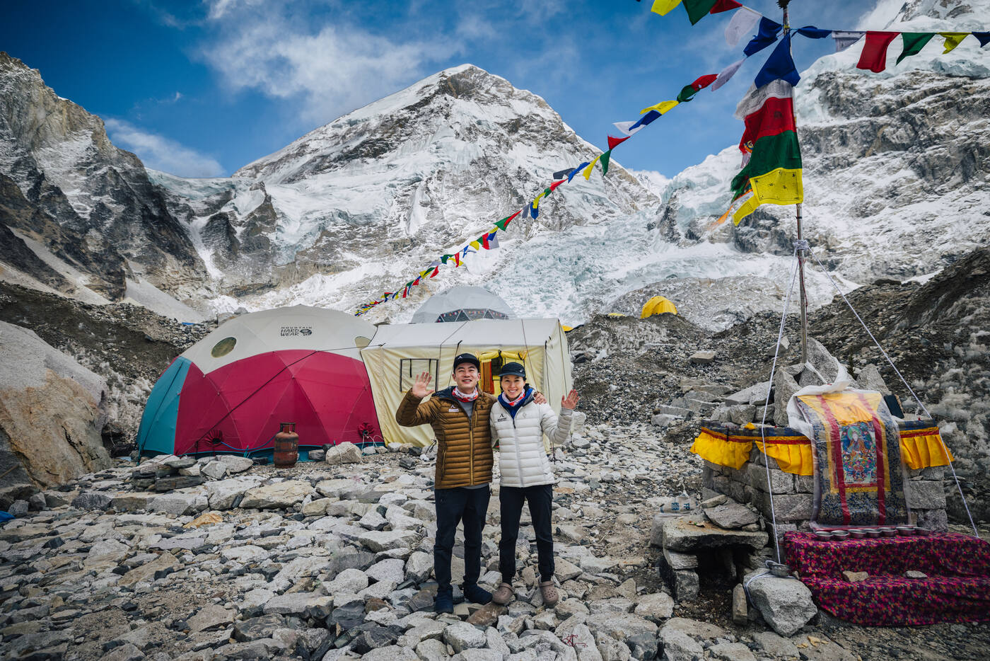 Two climbers waving in front of dome tents with prayer flags and the Puja altar at Everest Base Camp
