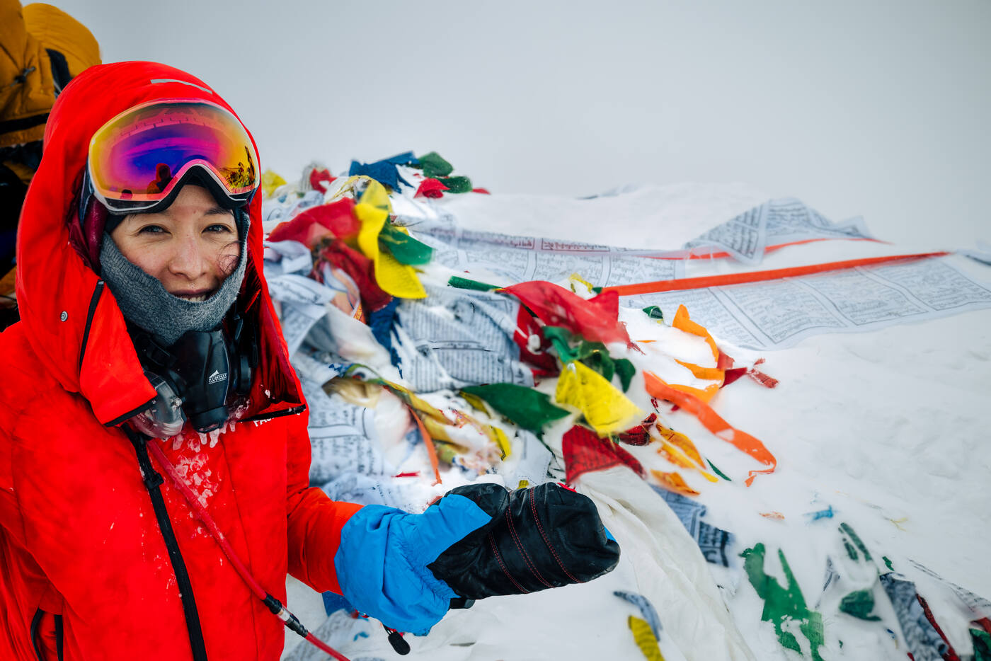 Climber in red jacket at the summit of Everest surrounded by prayer flags and snow